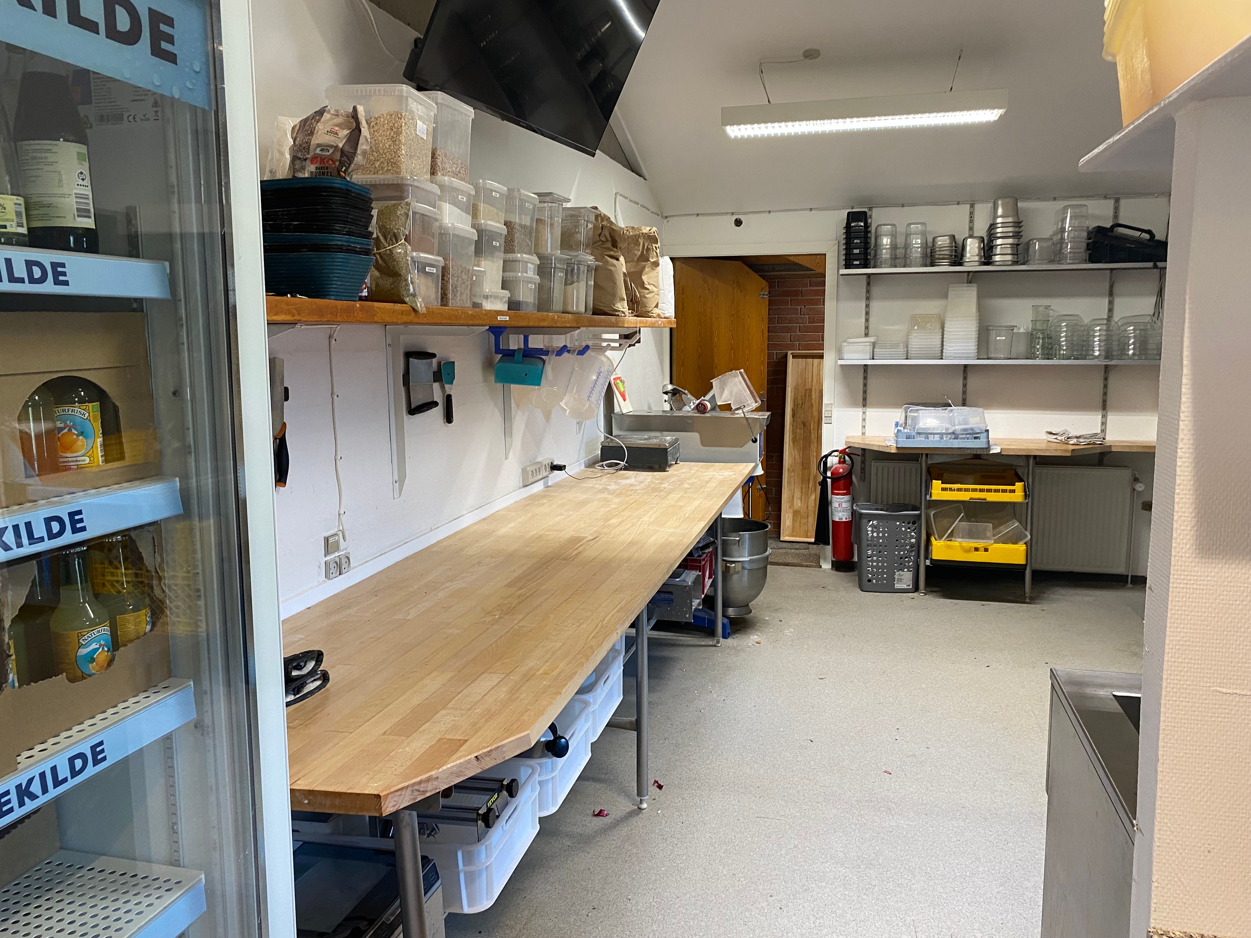 Køkken med træbord, krydsfinerhyller, ølflasker i køleskab, køkkenredskaber, plastbeholdere, glas, rør, slanger, sladre,