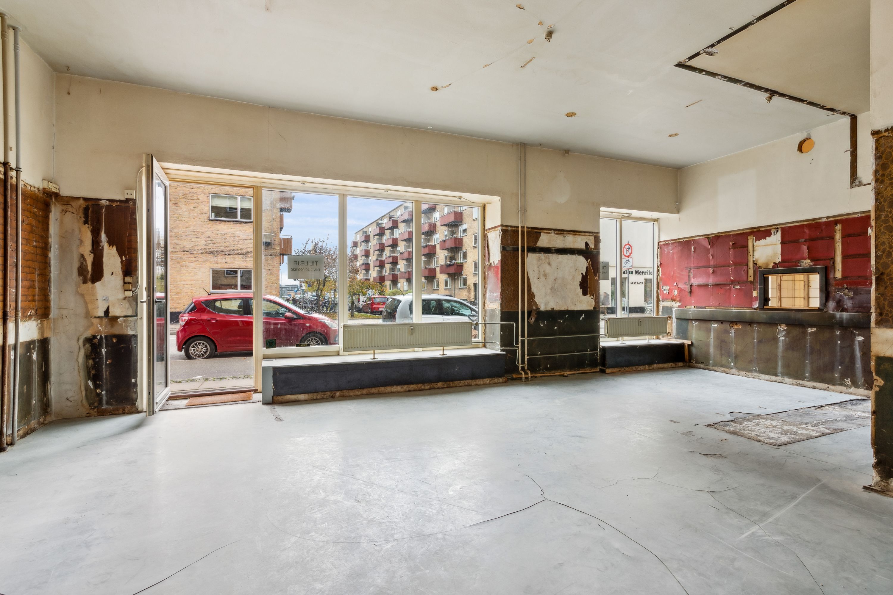 En tom lejemål med åben dør og røde vægge, hvor en bil er parkeret udenfor, og der er en høj bygning med røde altaner i