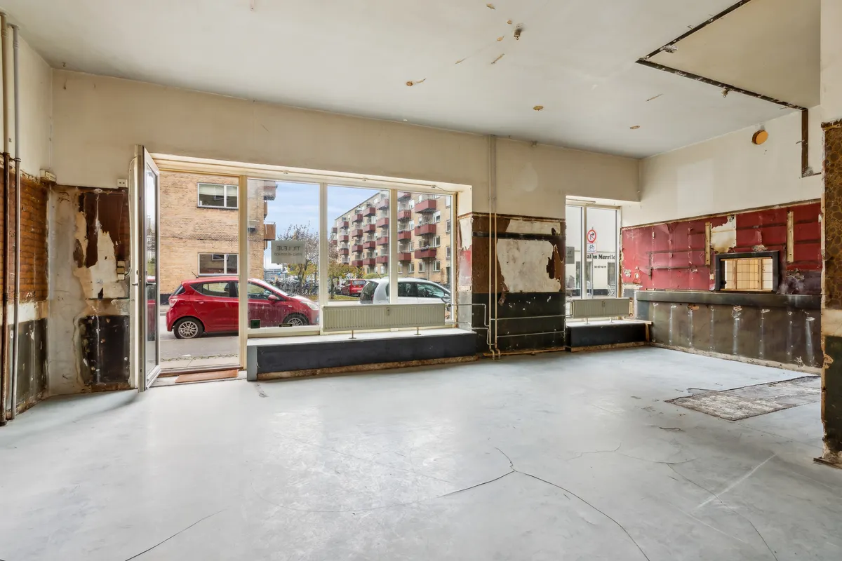 En tom lejemål med åben dør og røde vægge, hvor en bil er parkeret udenfor, og der er en høj bygning med røde altaner i