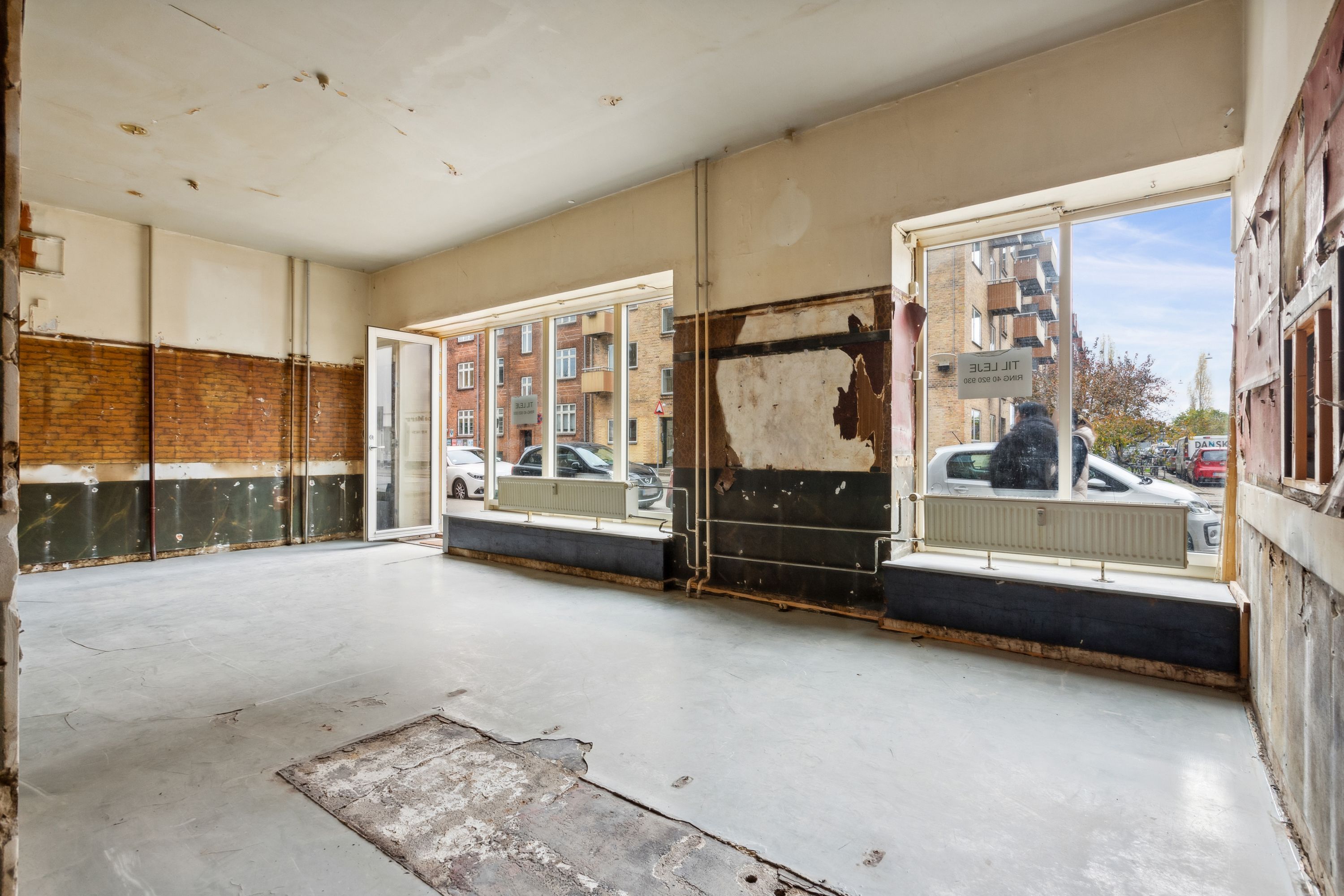 Et stort og rummeligt lokale med store ruder og en åben dør. En person står inde ved rudernes glas. Der er røde og brune