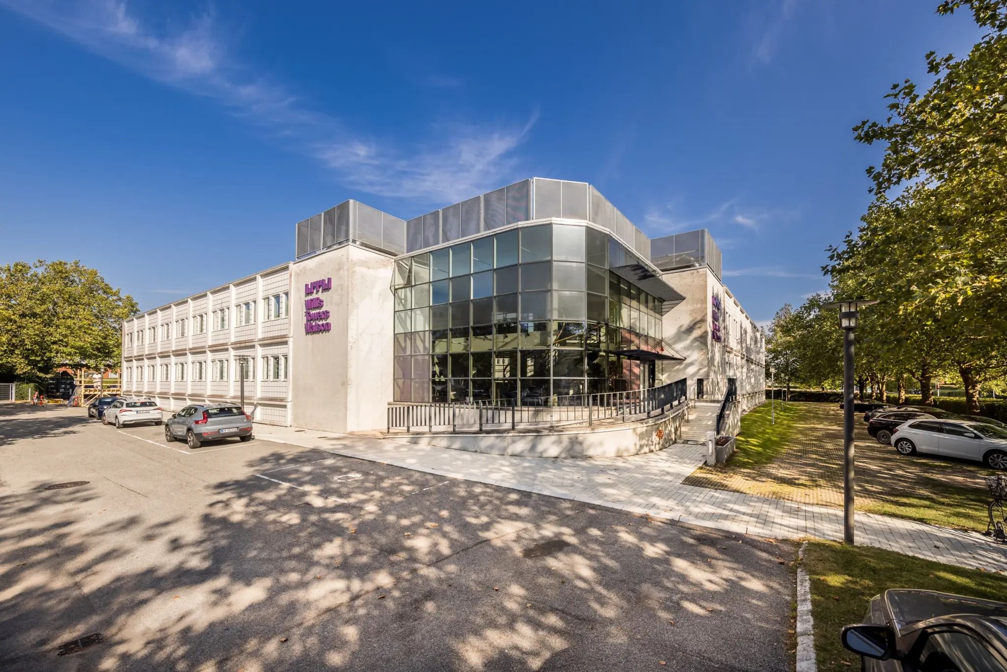 En stor bygning med glasvinduer og moderne arkitektur ligger ved siden af en park. Foran er parkeringspladser med flere