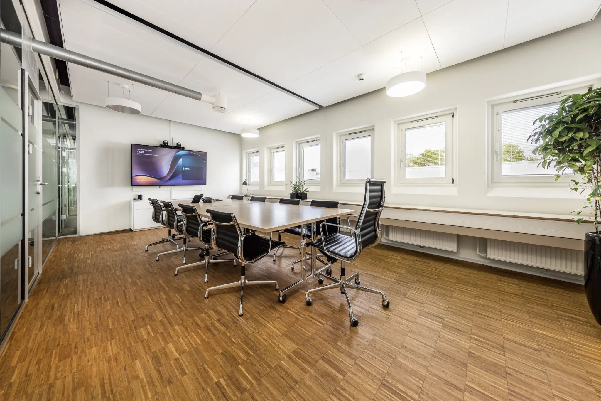 Mødeværelse med glasvinduer og hvide vægge, trægulv, og en stor tv-skærm samt to små planter