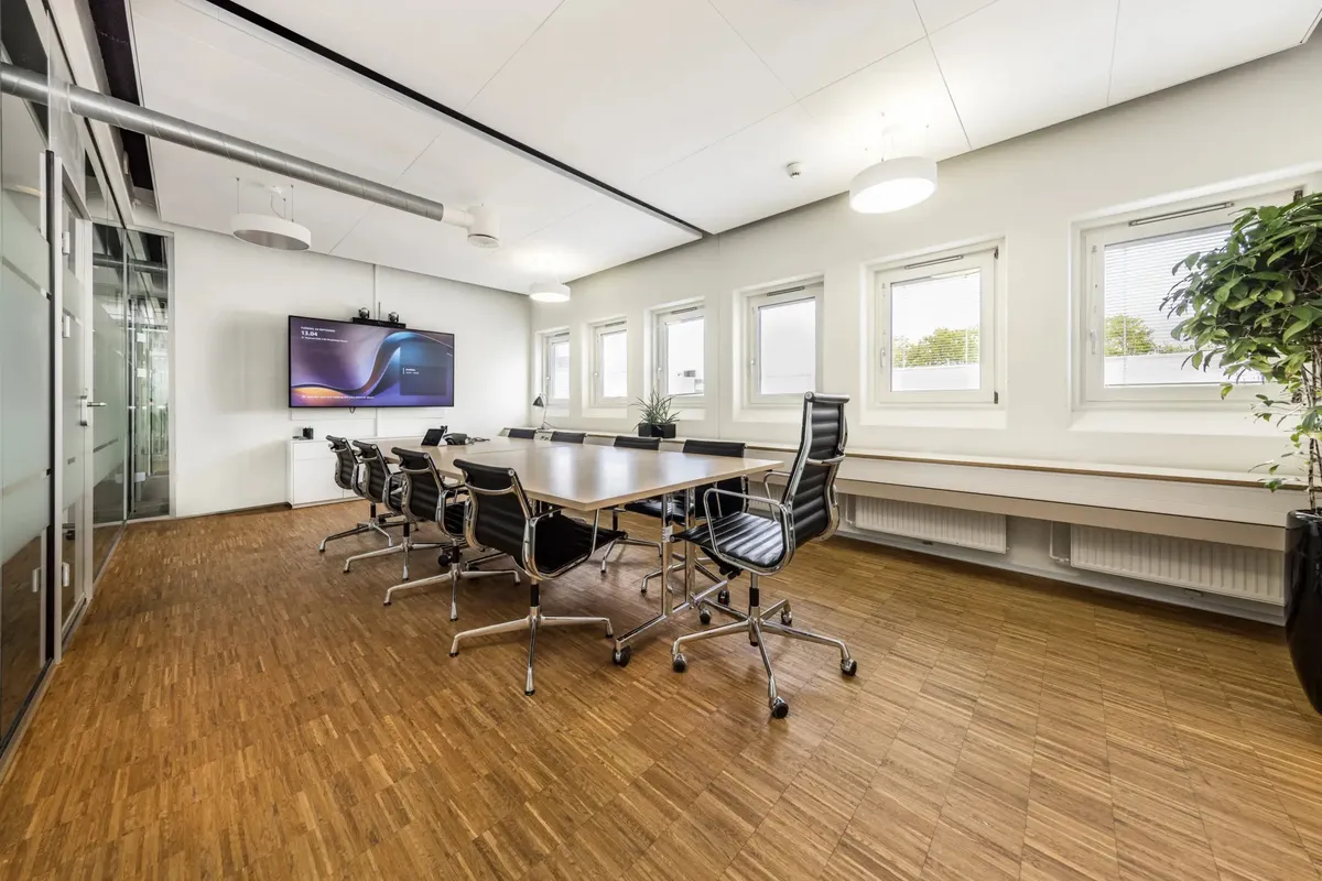 Mødeværelse med glasvinduer og hvide vægge, trægulv, og en stor tv-skærm samt to små planter