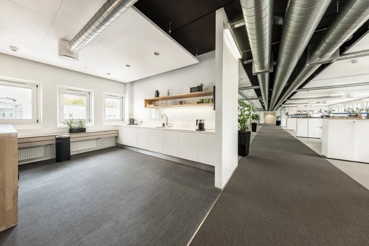 Et moderne kontor med hvide vægge og loft, gulvtæppe og højt loft med metalrør. Mange vinduer og et køkken med både en