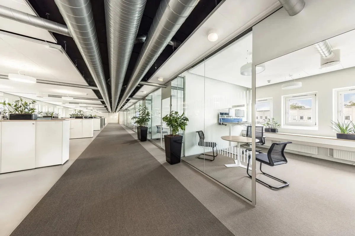 En moderne og veludstyret kontorgang med glasvægge og loftsventilation samt bord, stole, stolpeplanter og lysekroner