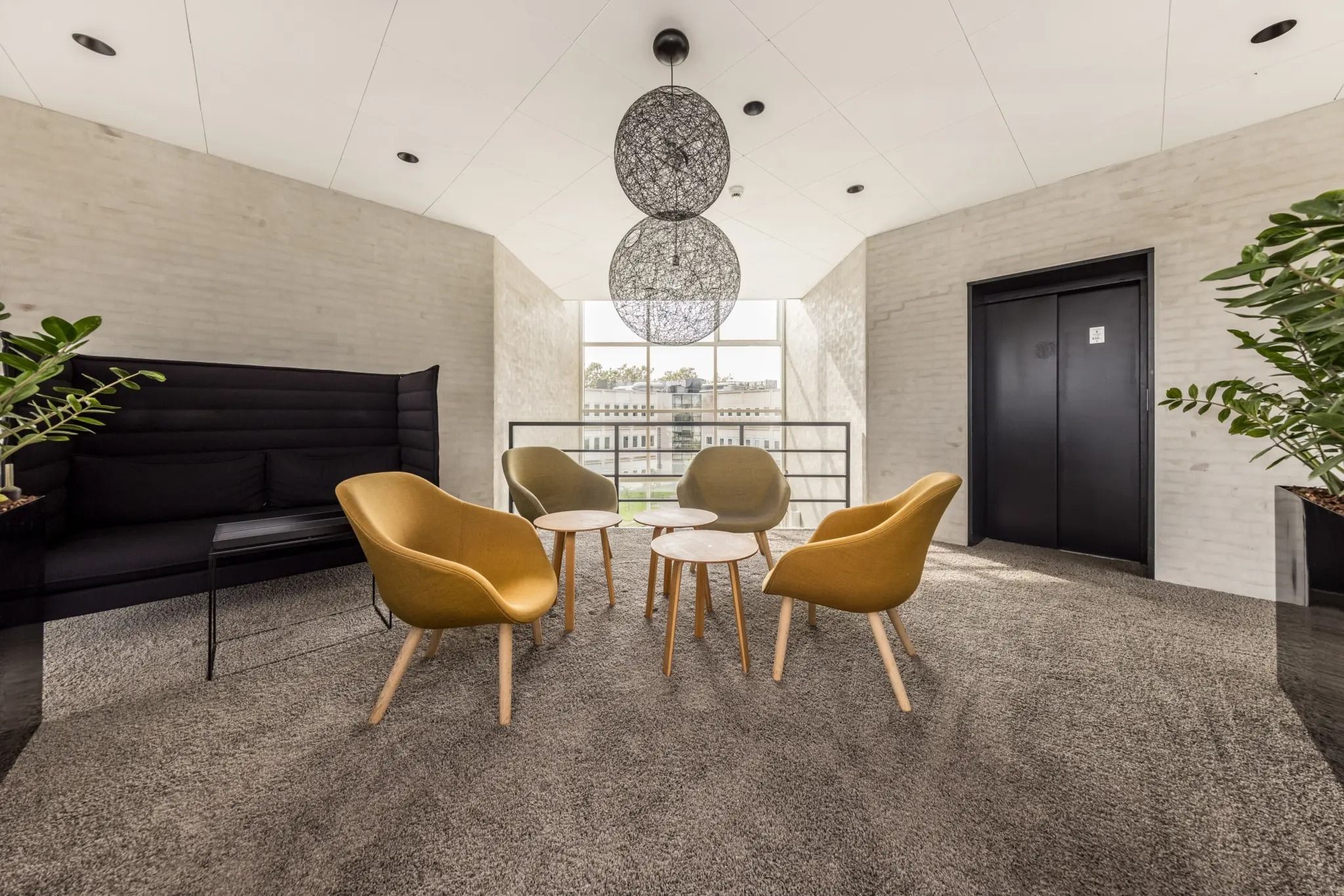 Indenfor er der et moderne lobbyområde med beige gulv og hvide vægge, møbler i lysebrune farver og to store plantepotter i