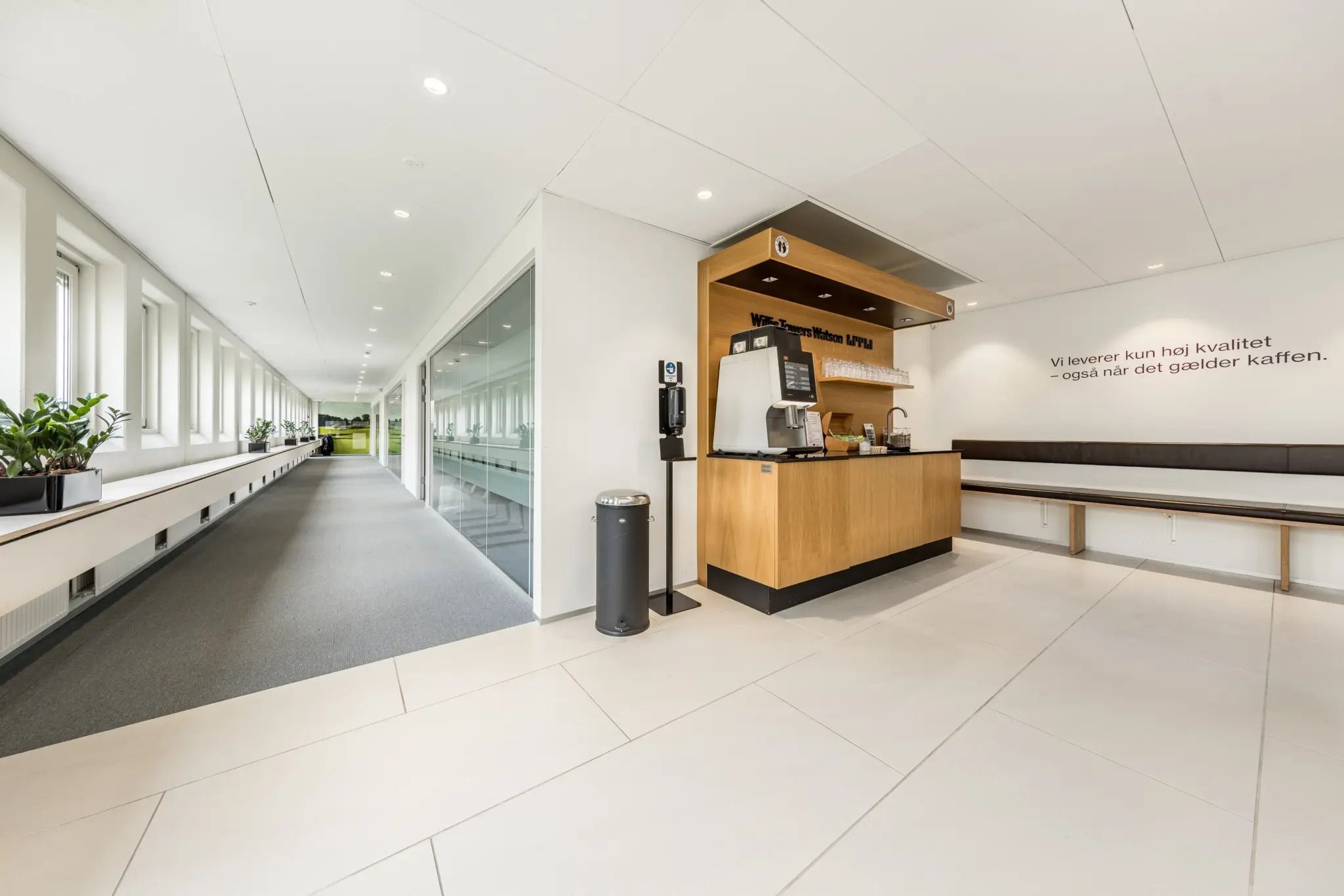 En moderne lobby med hvid gulvbelægning, en plante, en kaffemaskine og en tekst på væggen med teksten "Vi leverer kun høj