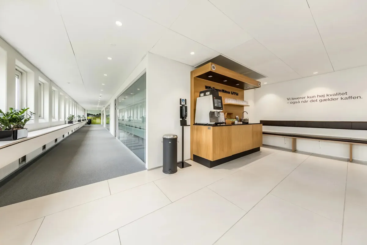 En moderne lobby med hvid gulvbelægning, en plante, en kaffemaskine og en tekst på væggen med teksten "Vi leverer kun høj