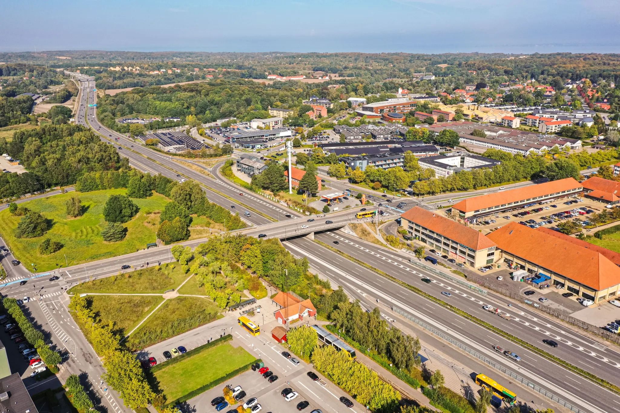 En luftfotooptagelse af Rundforbivej i Nærum med motorvej, parkeringspladser, bygninger, busser og grønne områder.