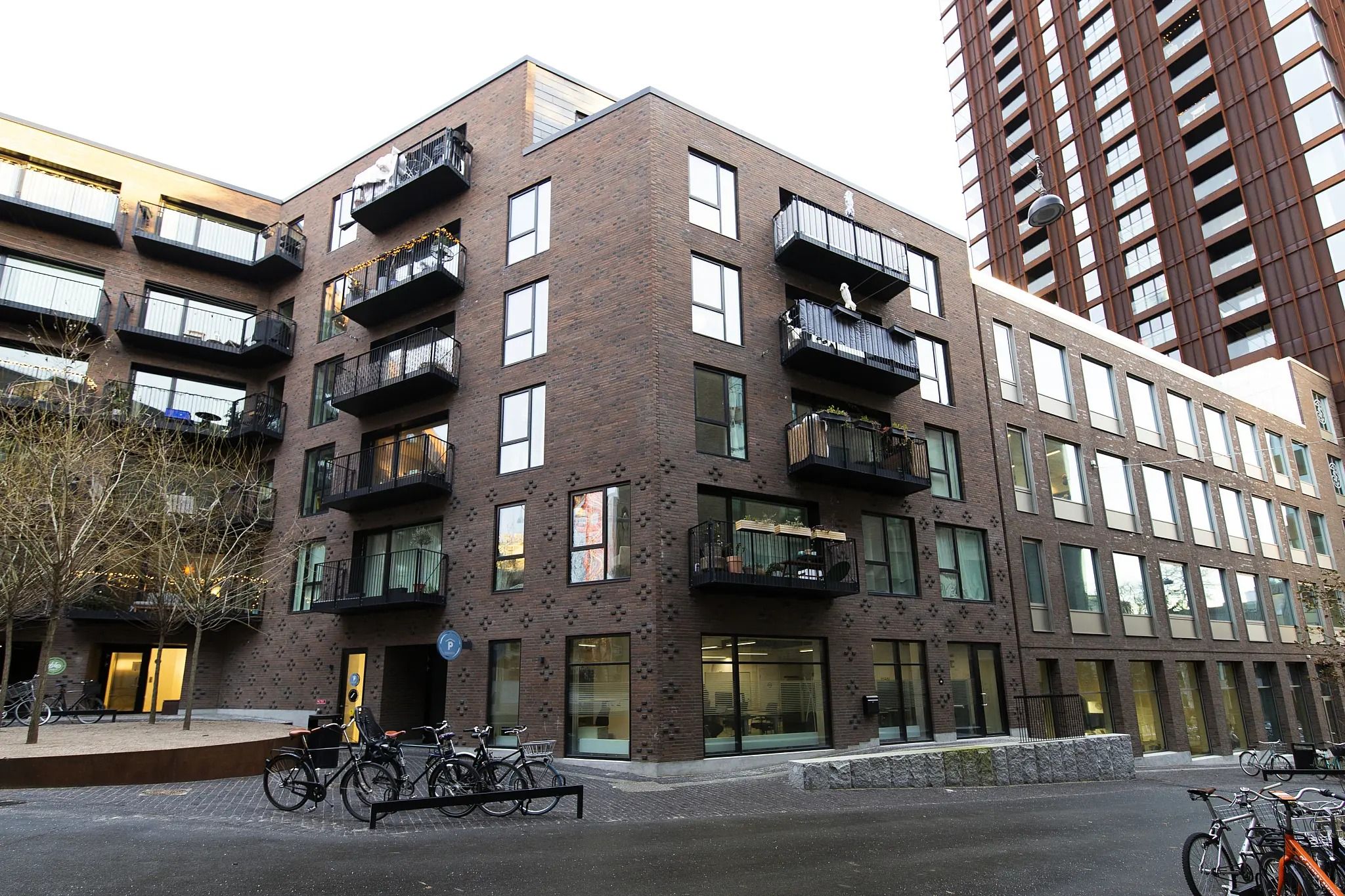 Bygning med terrasser og balkoner, fuld af cykler og plantager. Gågade med røde mursten og glasvinduer. Moderne design med