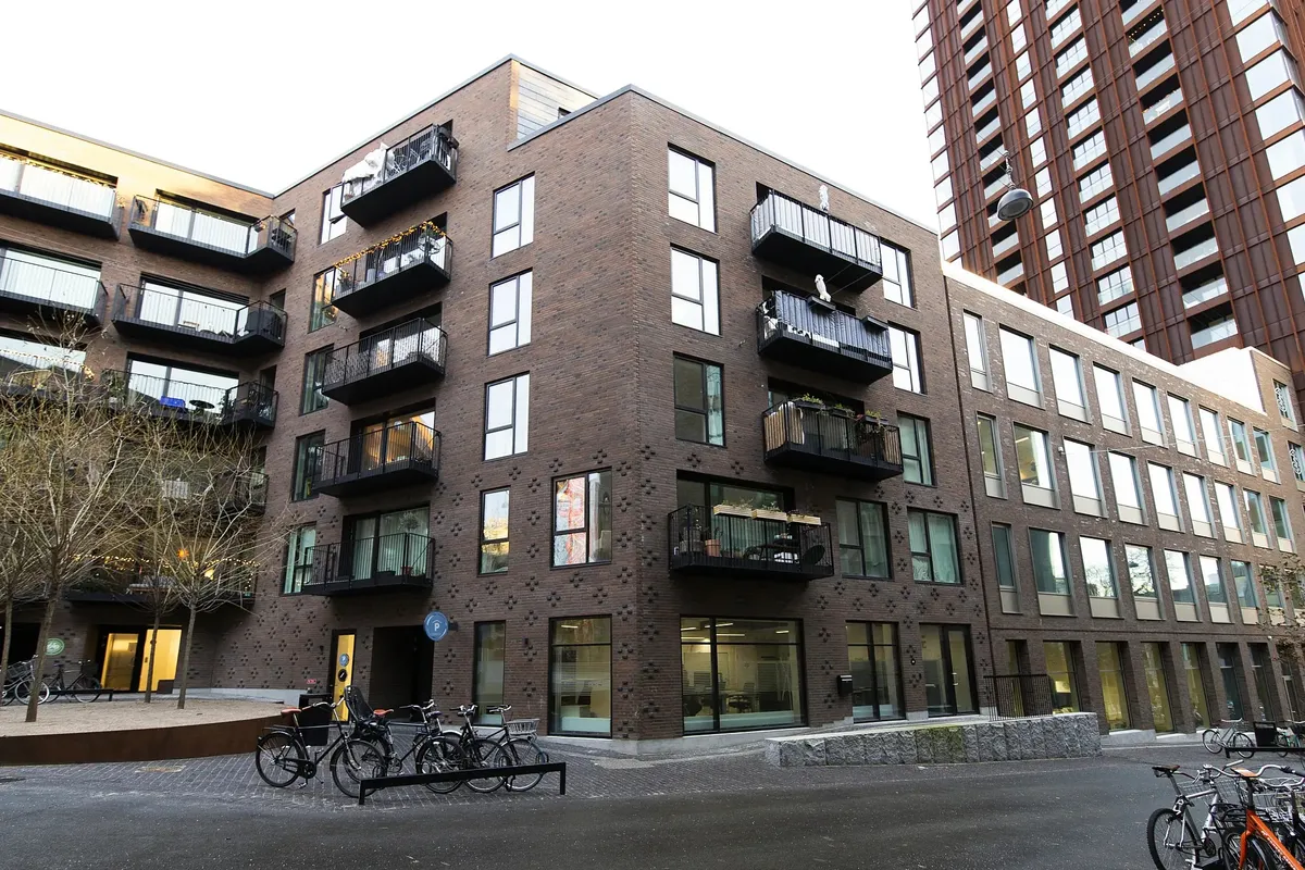 Bygning med terrasser og balkoner, fuld af cykler og plantager. Gågade med røde mursten og glasvinduer. Moderne design med