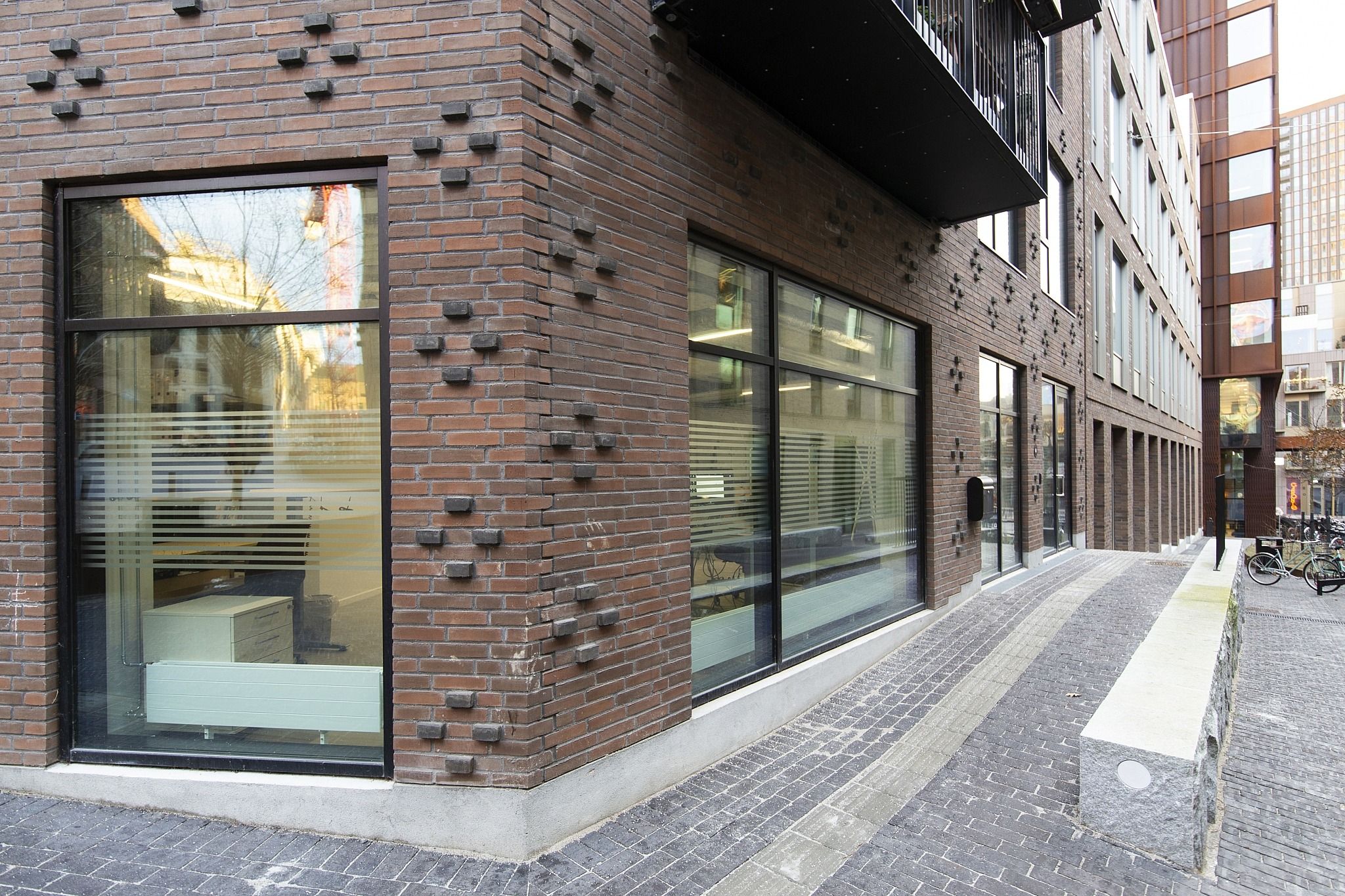 En ny bygning med glasruder og mursten. En række kontorer og et parkerede cykler langs gaden. Et skarpt og funktionelt