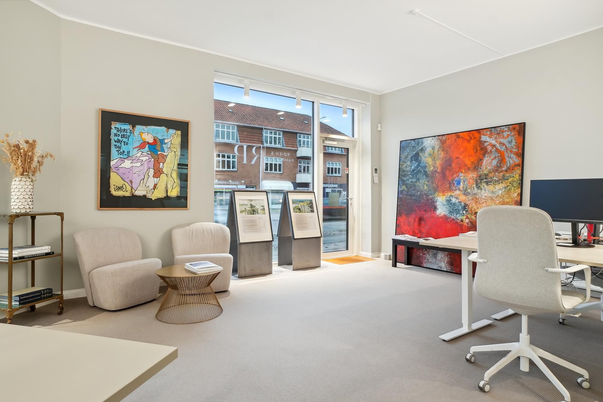 Inde i et kontorlokal er der to hvide lænestol, et bord med en computer, og et stort maleri på væggen