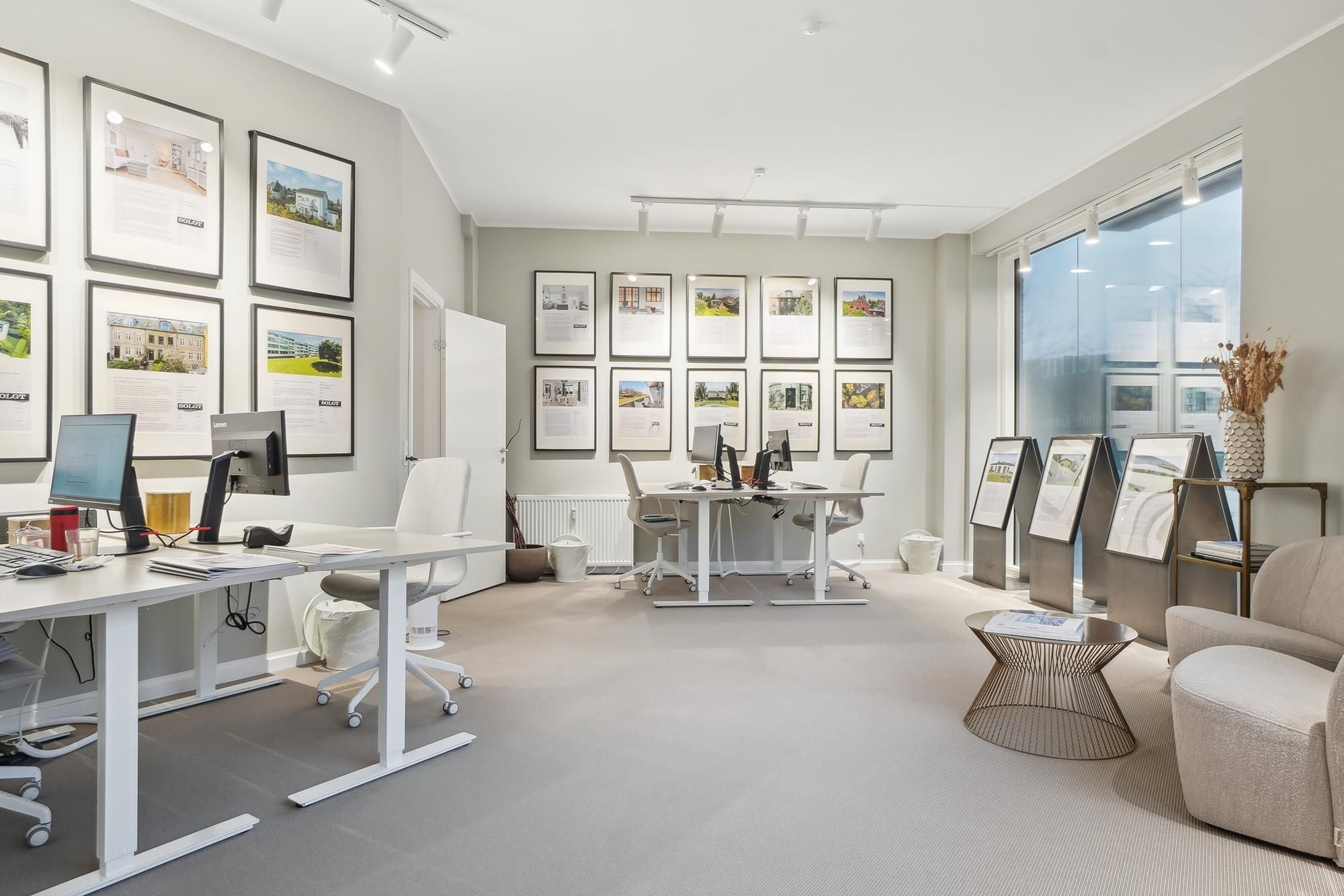 Kontor med højloft og store vinduer, hvide borde, lyse gulve, moderne møbler og fotorammer på væggen