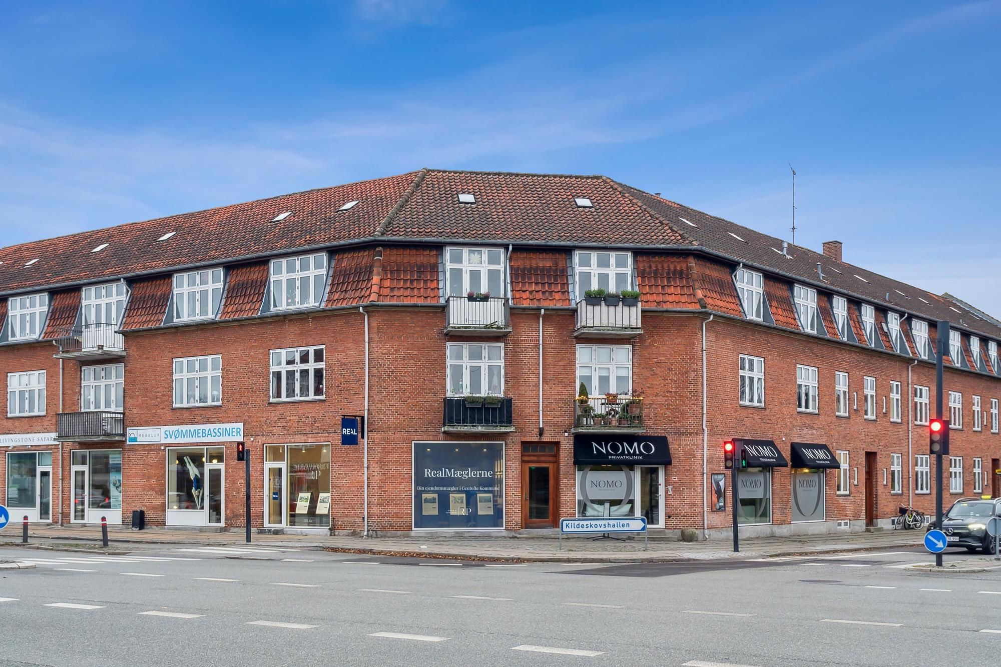 Et rødt bygningshus med flere butikker og vinduer, hvor en er udstillingsvindue til RealMaglerne
