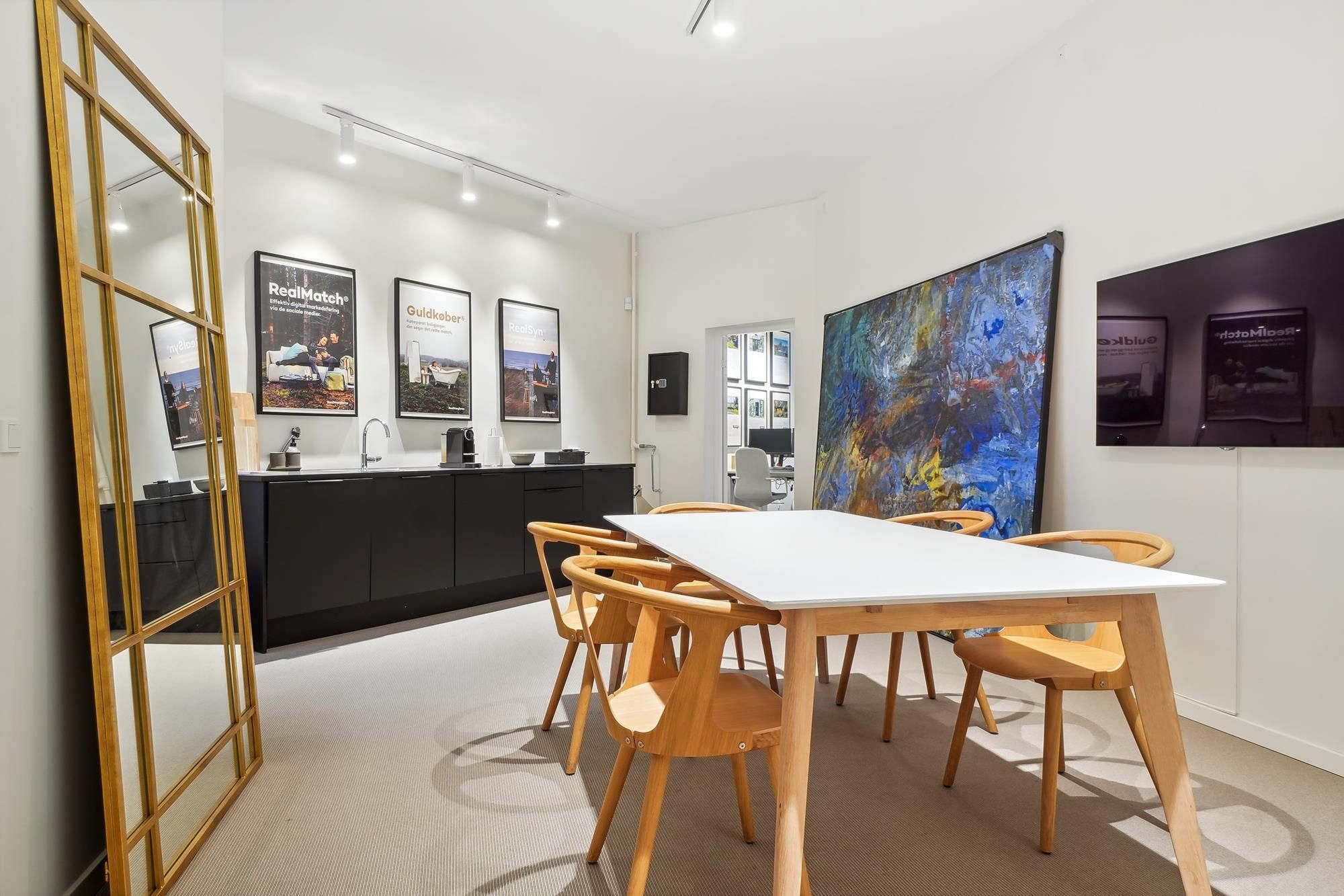 Kontorrum med højt loft, hvidt bord og lyse stolene, hængslet spejl og store kunstværker. En moderne køkkendeling med høj