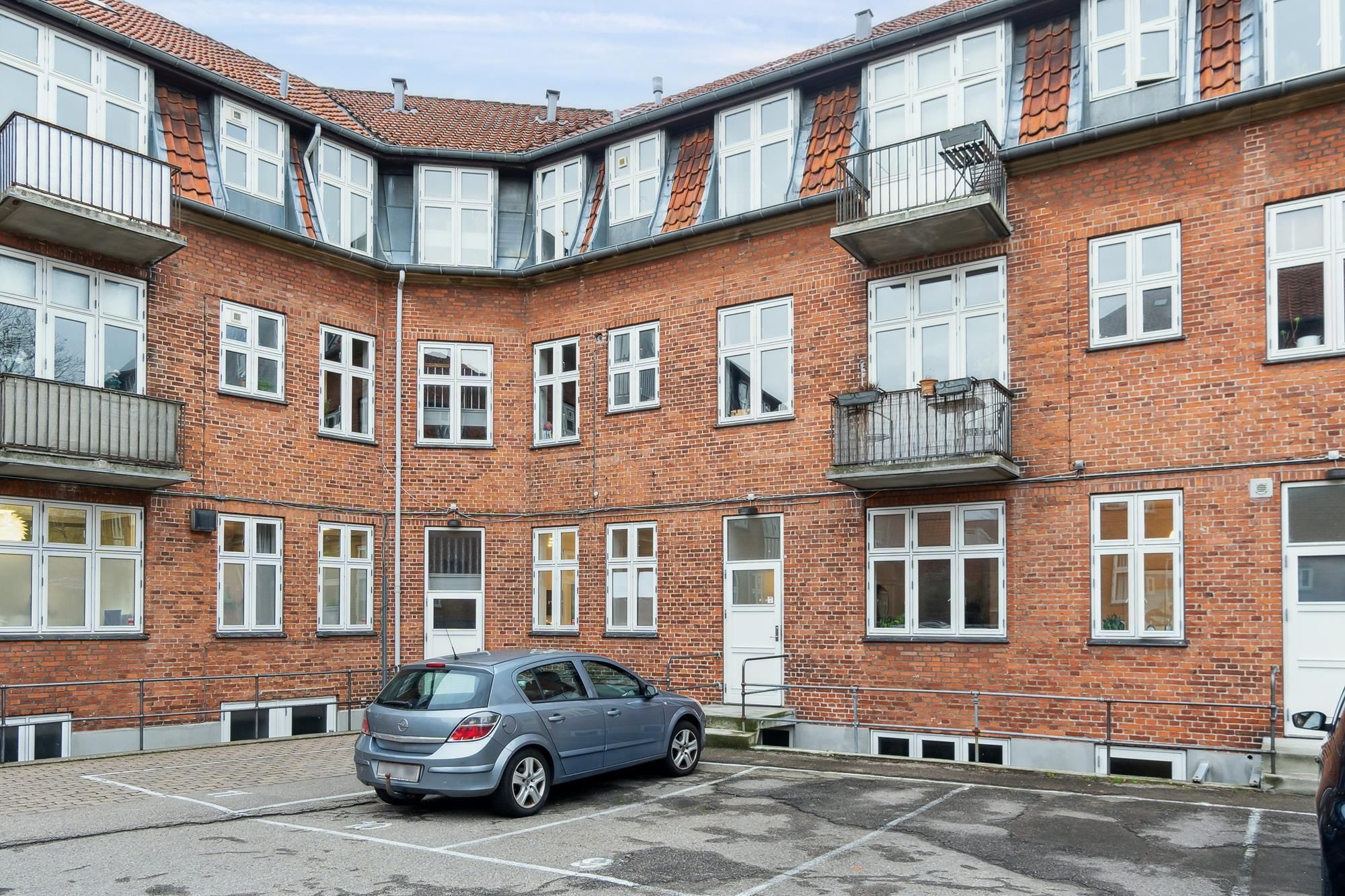 Det er en rødmalet lejlighedsbygning med fire etager og mange vinduer, og der er en bil parkeret på fortovet.