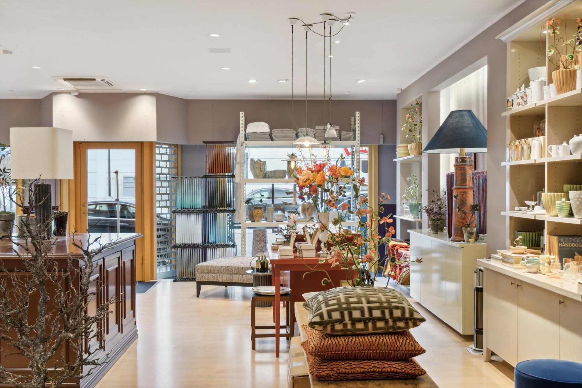 Lyst og rummeligt erhvervslejemål med lyse loftslamper, hylder og gulv, smukt indrettet med puder og blomster