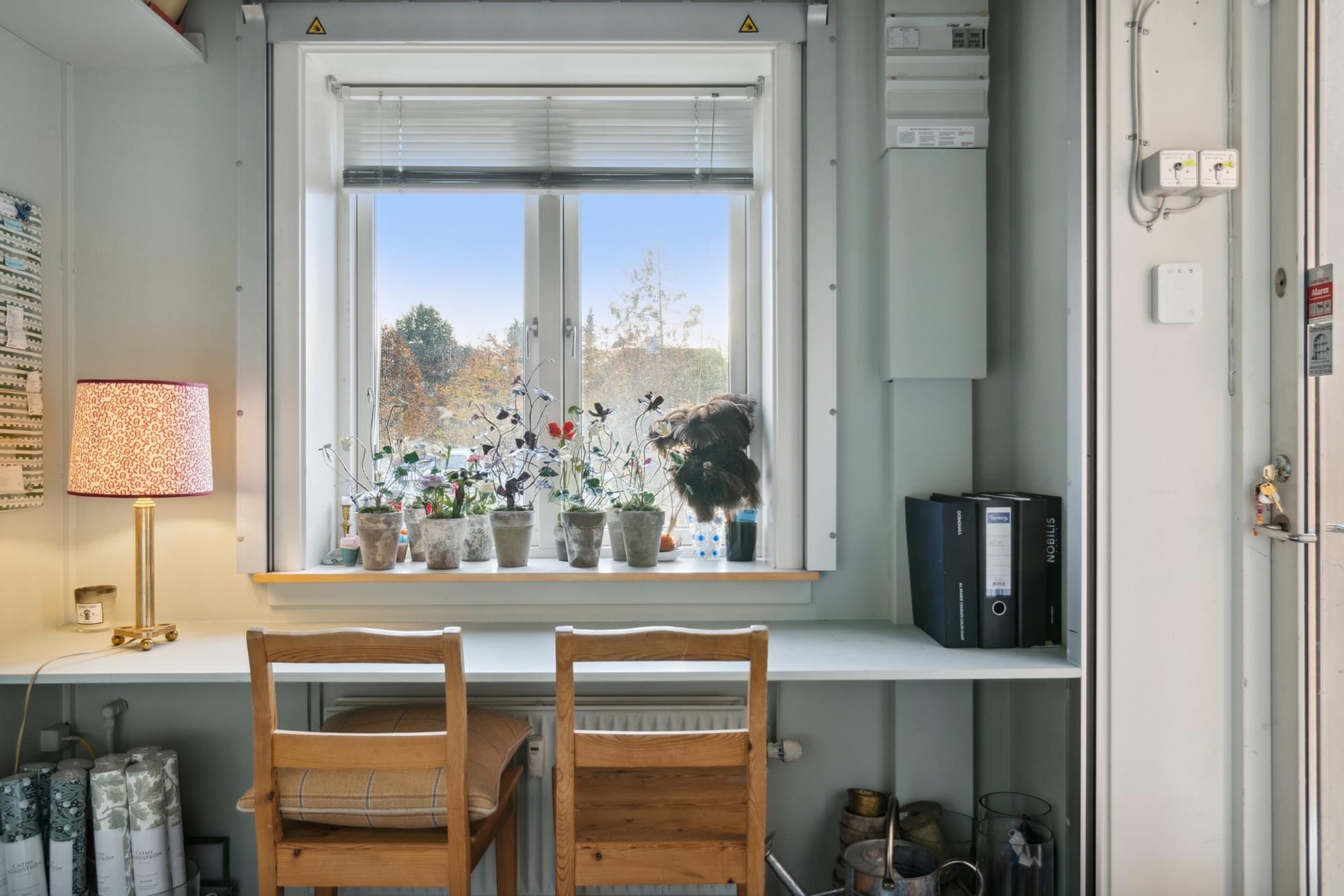 Åbent og lyst værelse med vinduer og plantepot, to stole, højtalere, lampe og et skab med møbler og filer