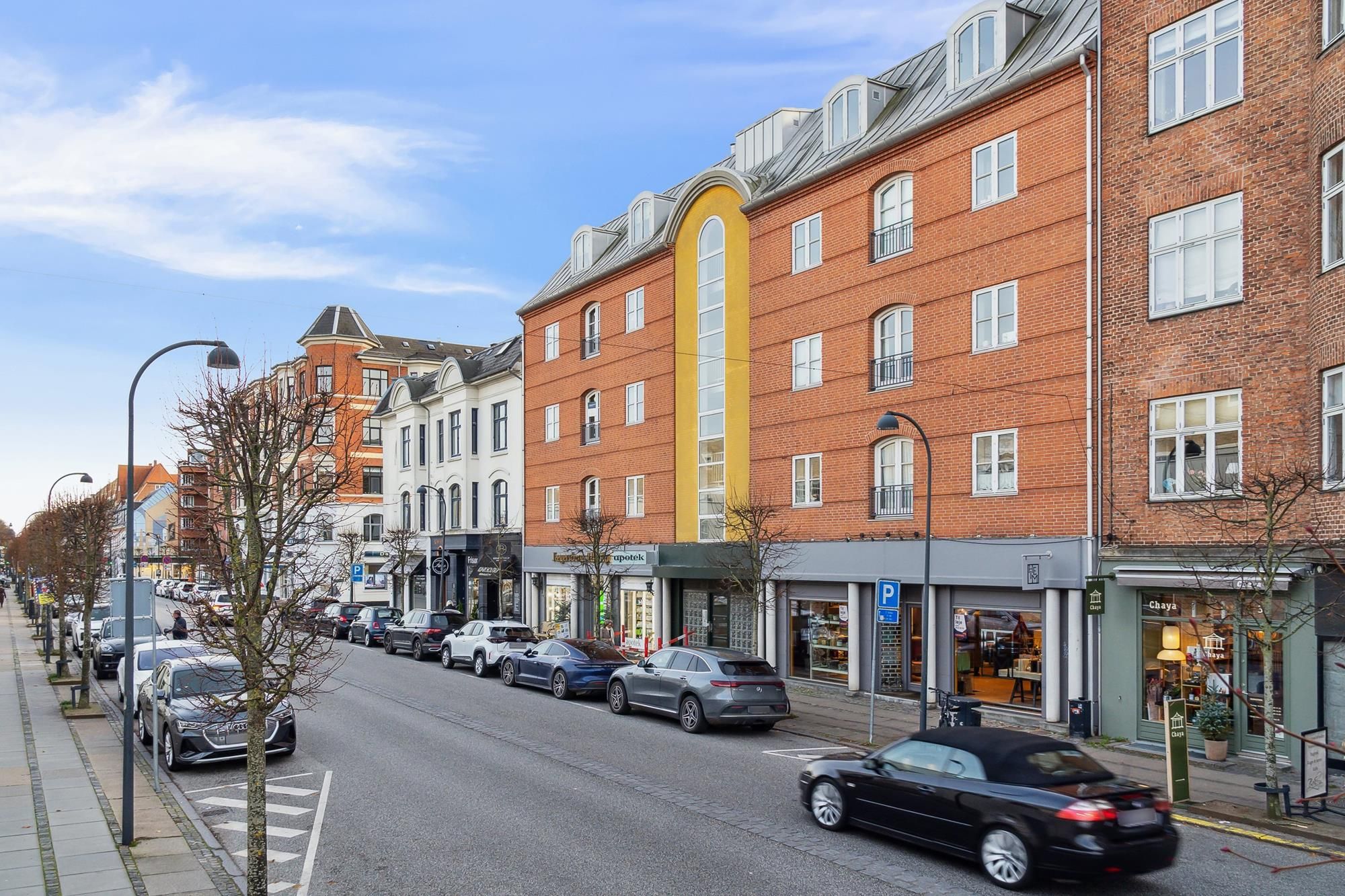 En bil kører ned ad en gågade med forretninger og parkerede biler, træer og gadelamper