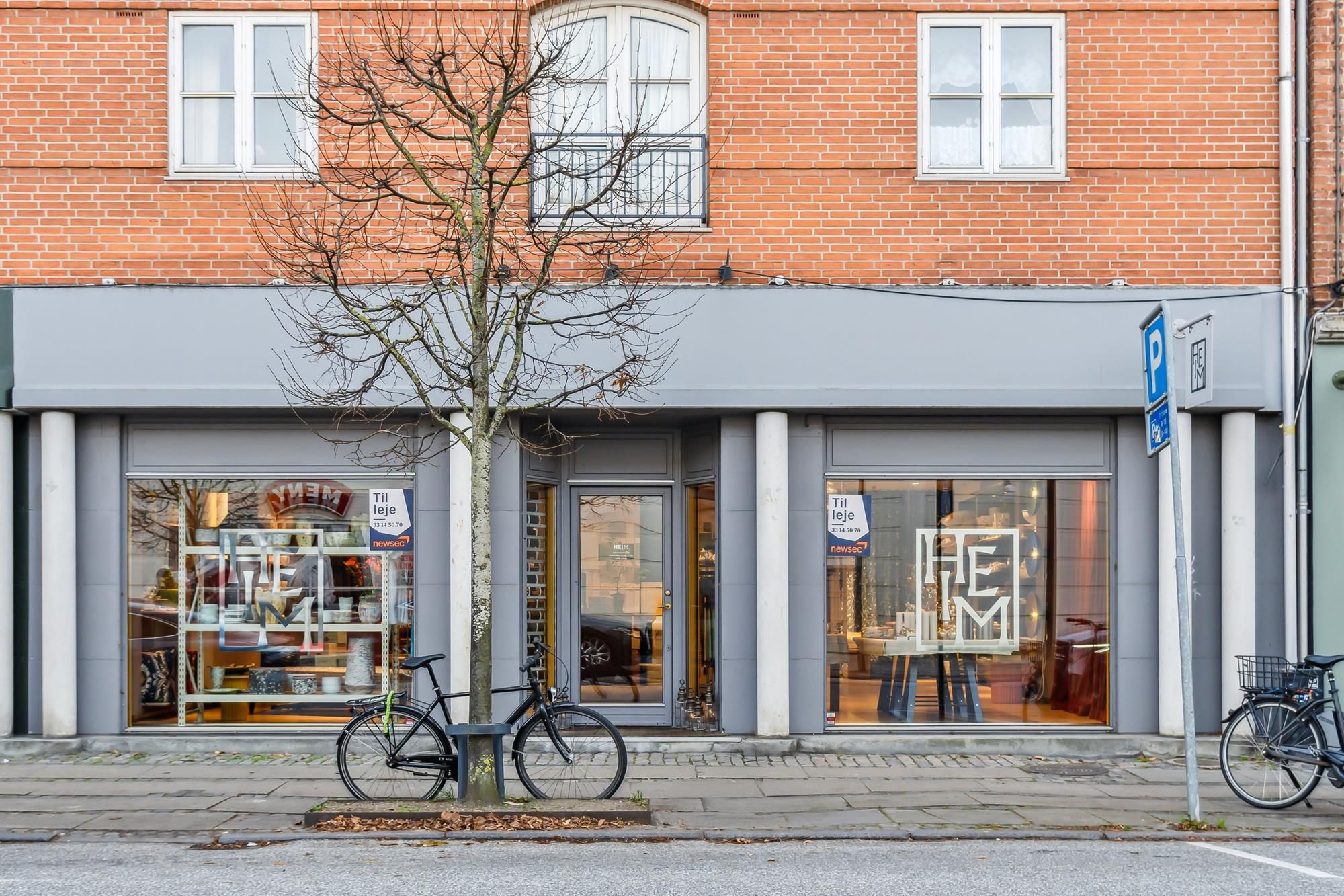 Erhvervslejemål med store glasvinduer og smukke facader, hvor der er to cykler parkeret på fortovet.