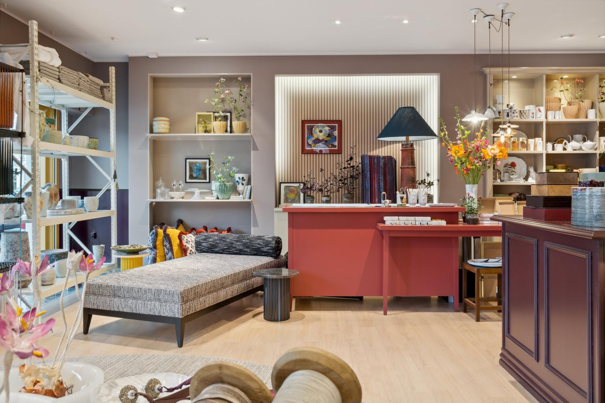 Dekorativt lokale med lyse vægge, hylder med tæpper og keramik samt et rødt bord og lysekroner