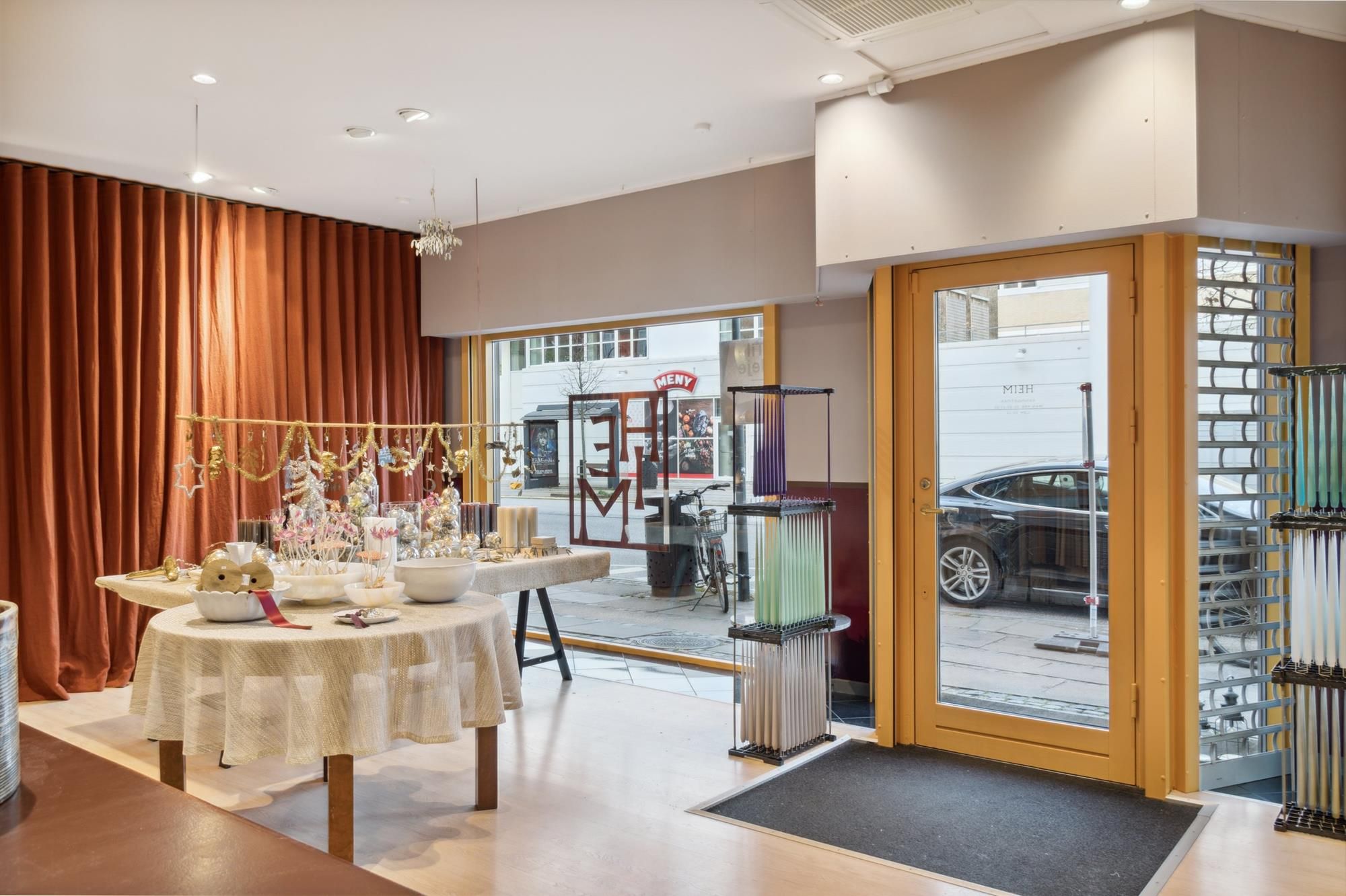 Lyst lokale med store vinduer, møbler, lysekroner og udsmykning samt udsigt til en bilparkeret på gaden og en møbelbutik i