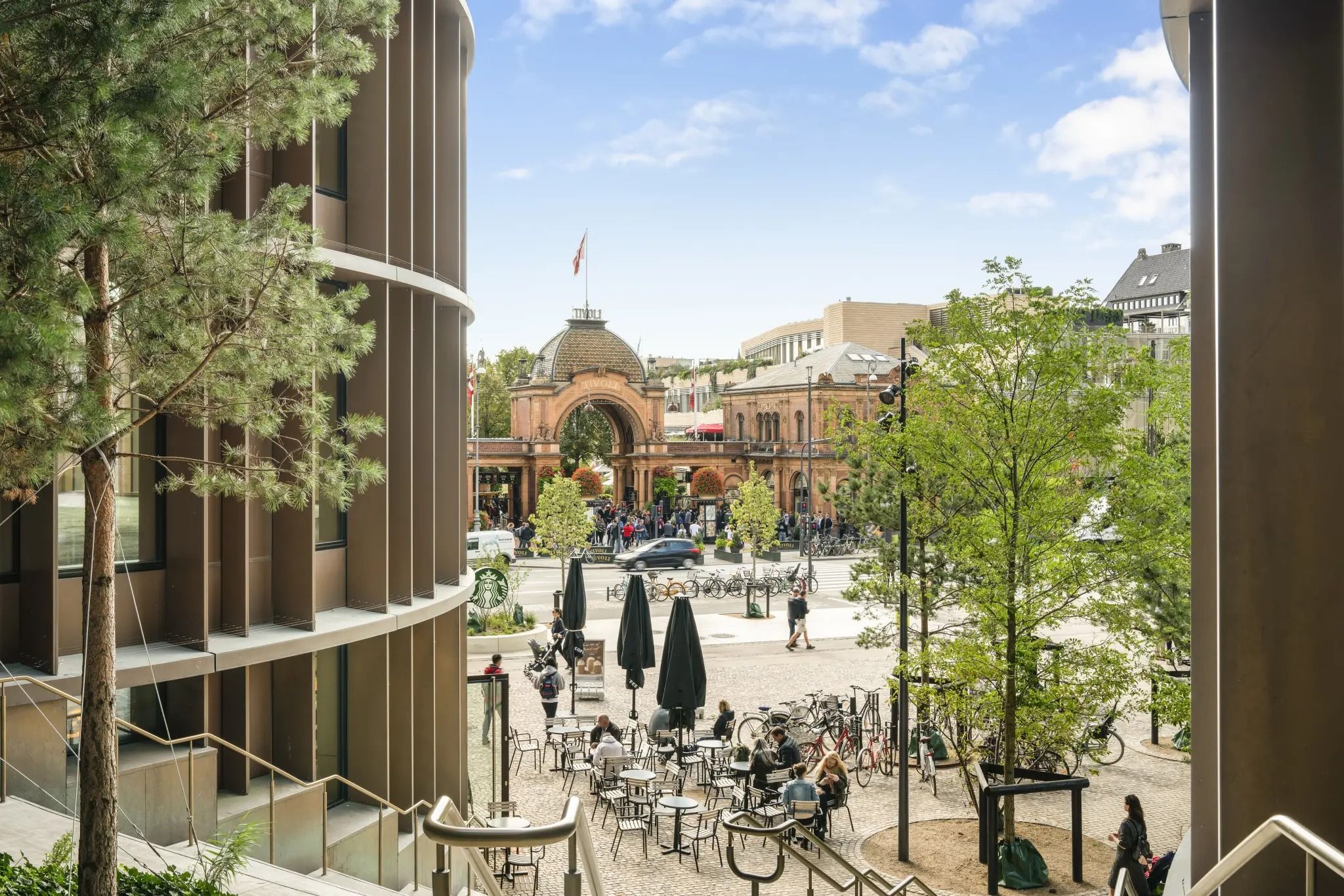 En gårdplads med træer og caféborde og stole uden for Tivolis indgang. Borgere går og står rundt, og nogle cykler forbi.
