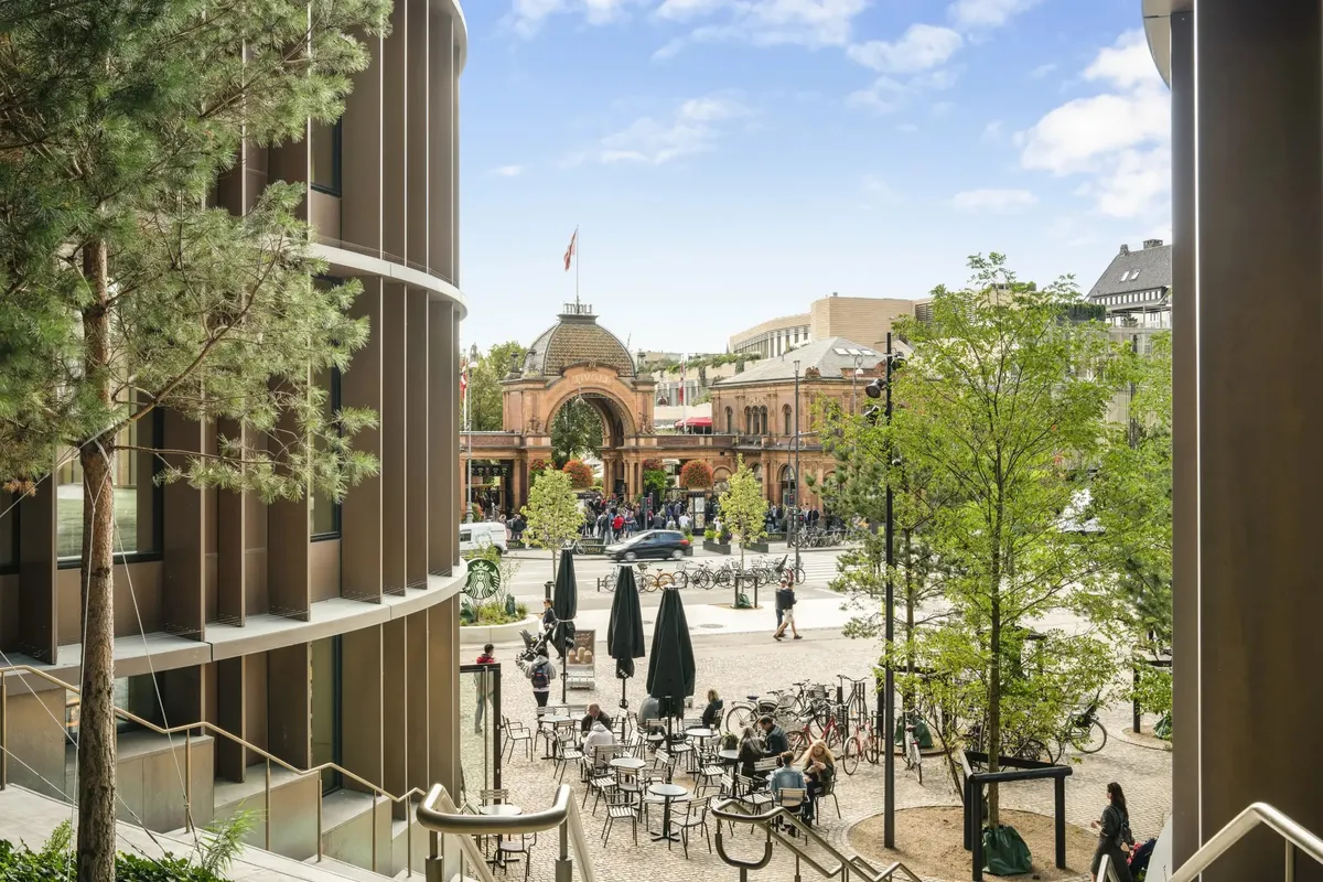 En gårdplads med træer og caféborde og stole uden for Tivolis indgang. Borgere går og står rundt, og nogle cykler forbi.