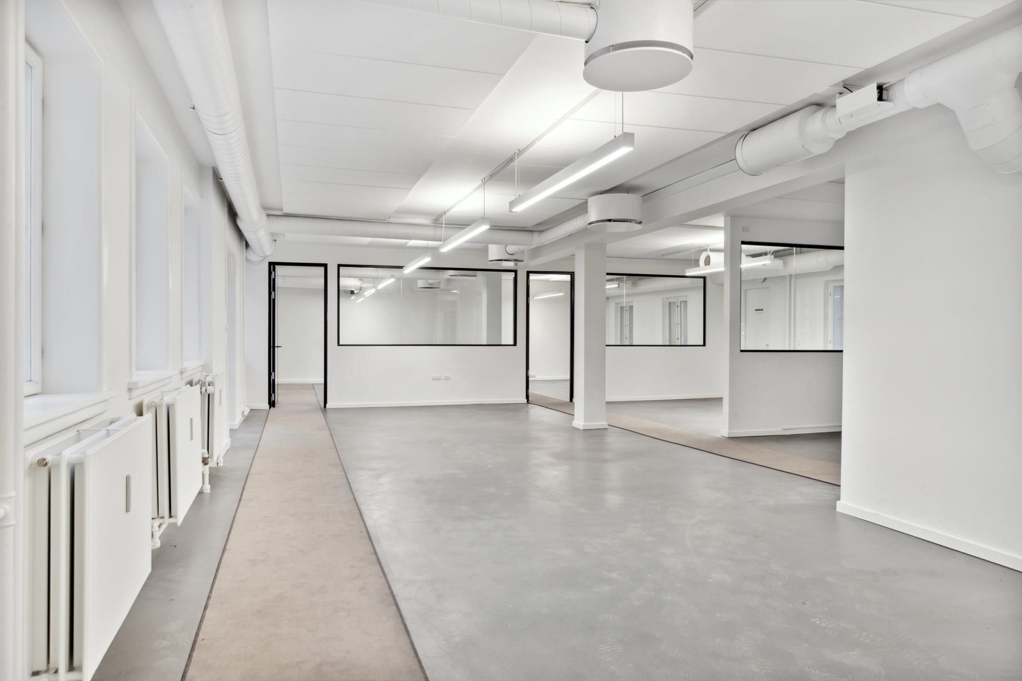 Kort korridor i moderne kontorbygning med hvide vægge, lysekroner og glasdøre samt lysegulve og radiatorer i hvidt