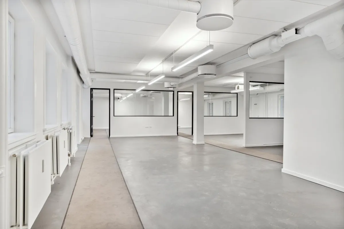 Kort korridor i moderne kontorbygning med hvide vægge, lysekroner og glasdøre samt lysegulve og radiatorer i hvidt