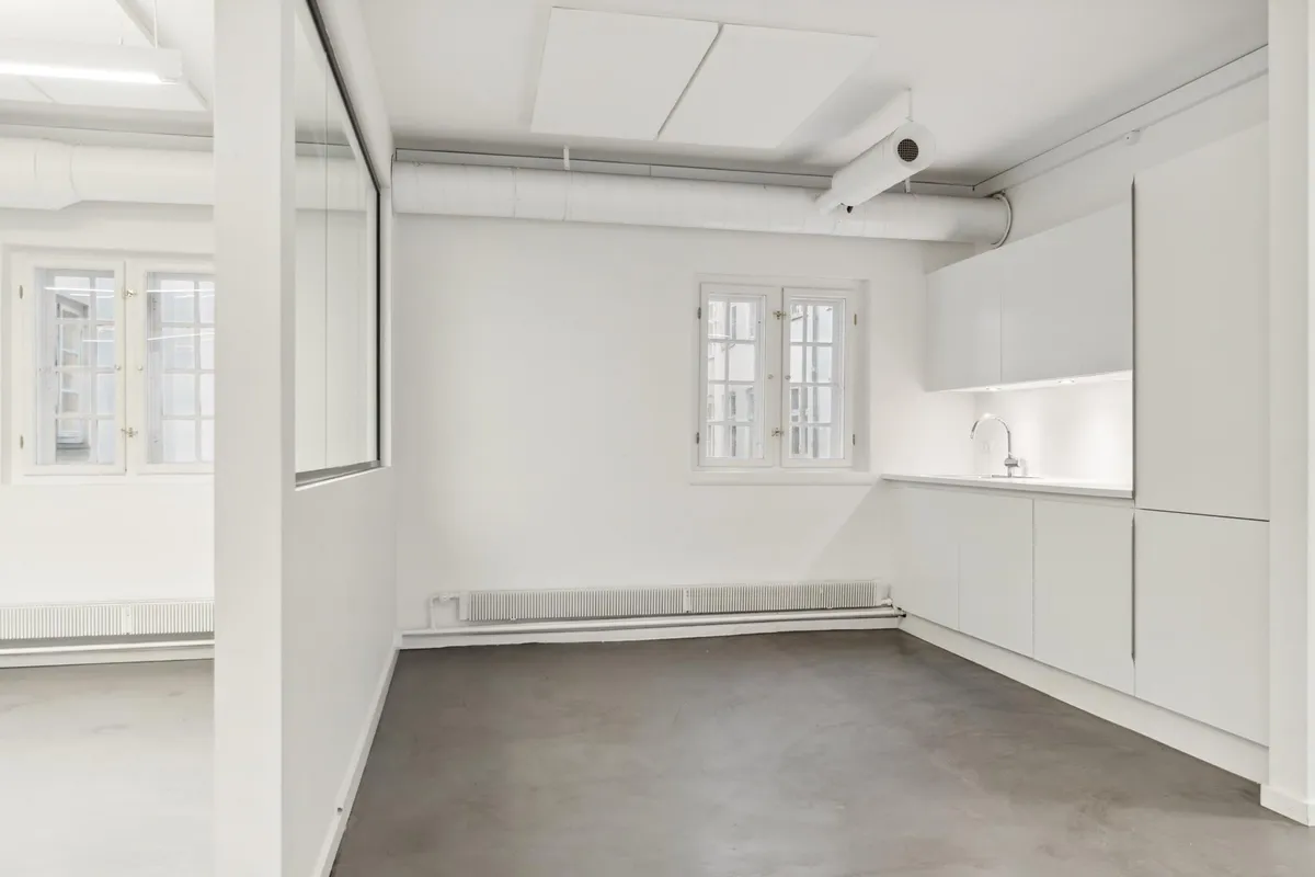 Et tomt kontor med hvide vægge og gulve, hvidt loft, en radiator og to vinduer med hvide ruder