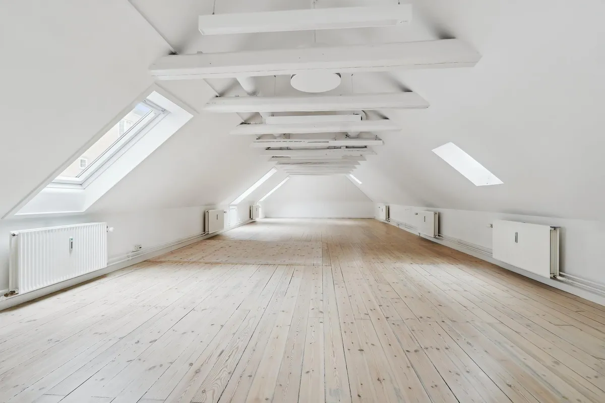 Hvid loft med bjælker og loftsvinduer i et stort rum med lyse trægulve og hvide vægge med radiatorer