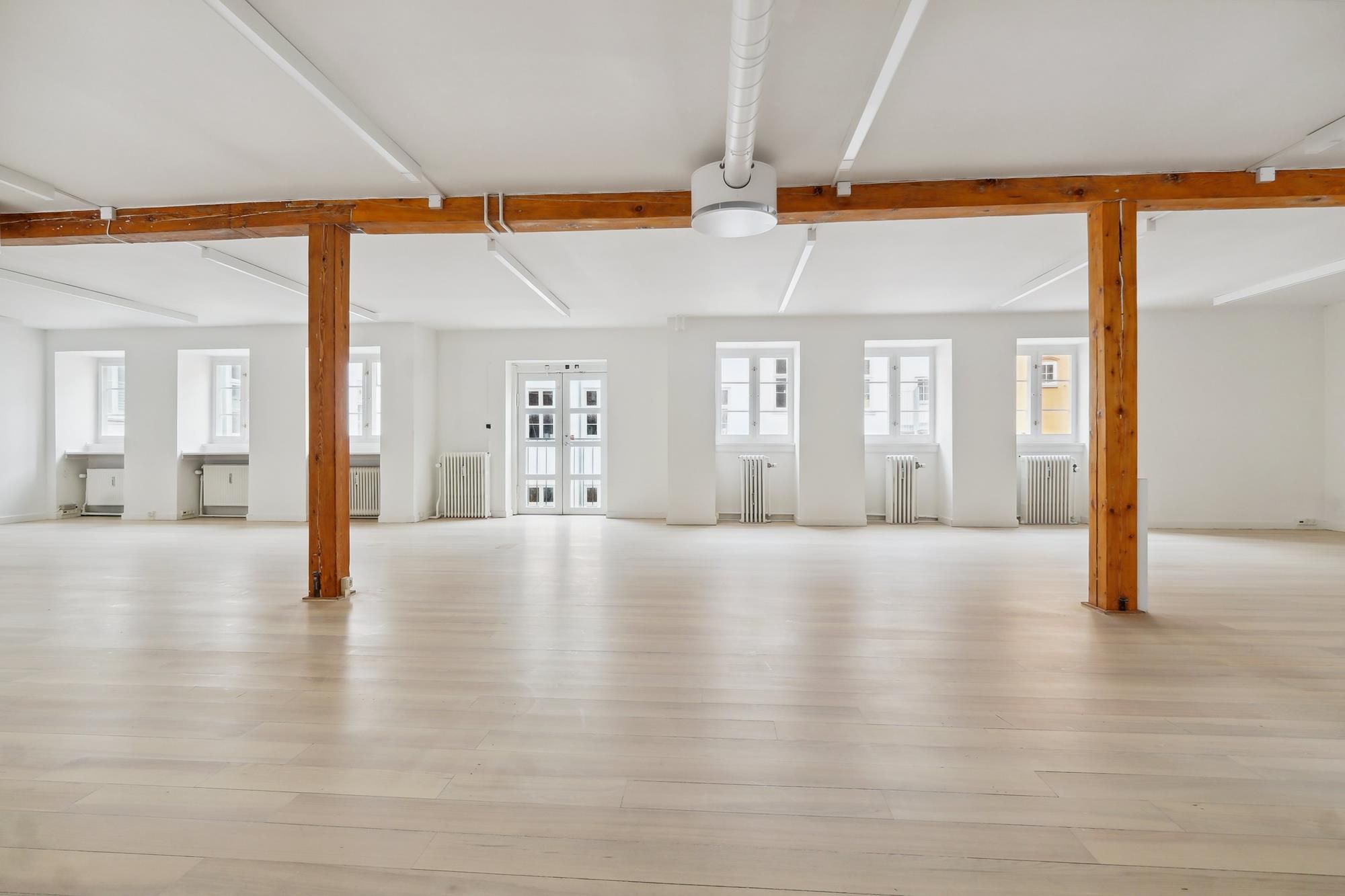 Elegant kontorlokaler med højt til loftet, hvidt loft, lyse gulvbelægning og to store vinduer i hver væg