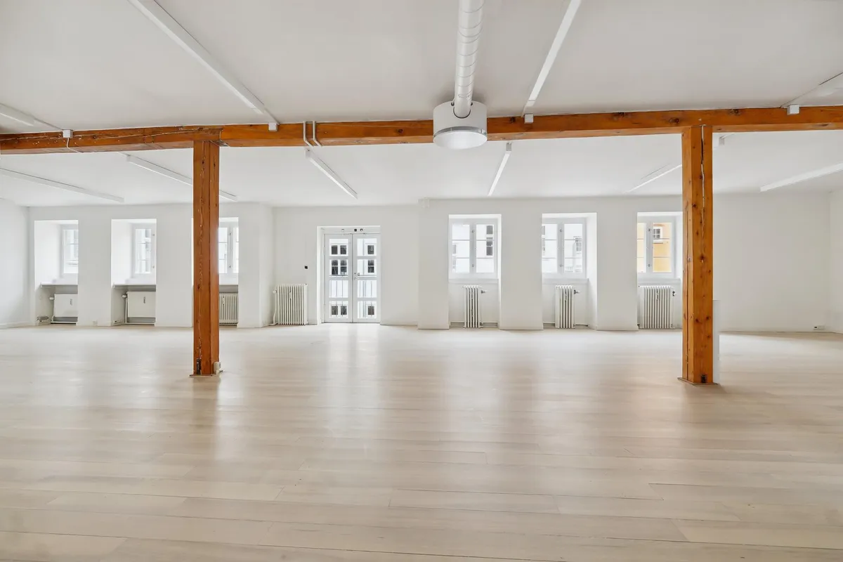 Elegant kontorlokaler med højt til loftet, hvidt loft, lyse gulvbelægning og to store vinduer i hver væg