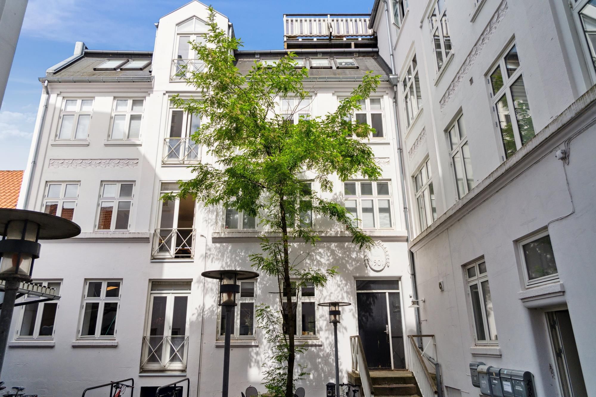 Hvid malet bygning med store vinduer, slanke gavlflader og en indgangsdør. En lille grøn træ med nogle bænke og to