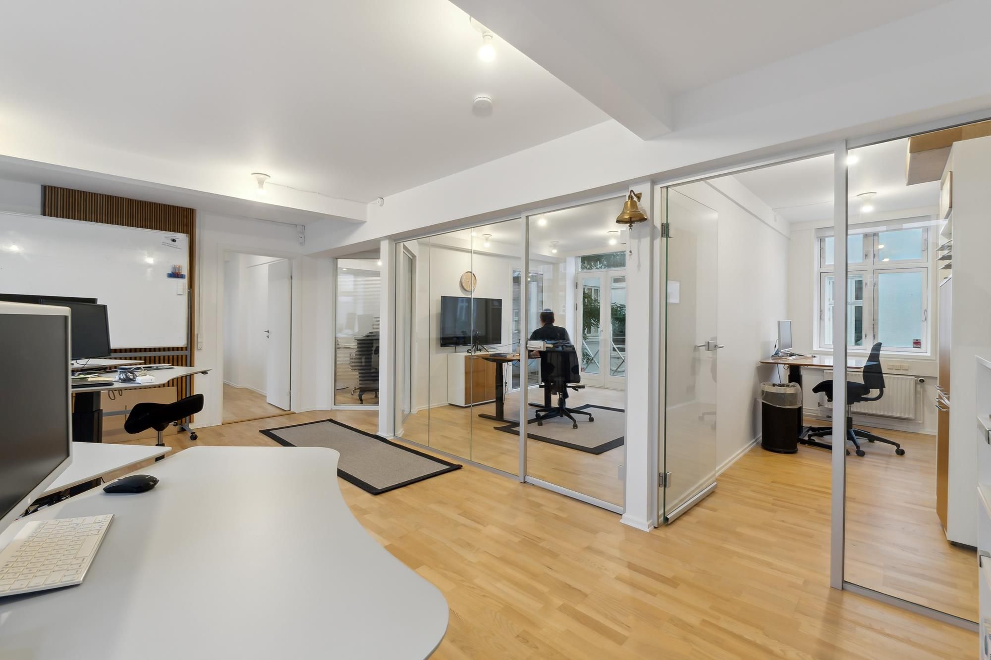 Åbent kontor med højt til loftet og lyse gulve. En person sidder ved et bord med computer og tv. Hjørne med en hvid