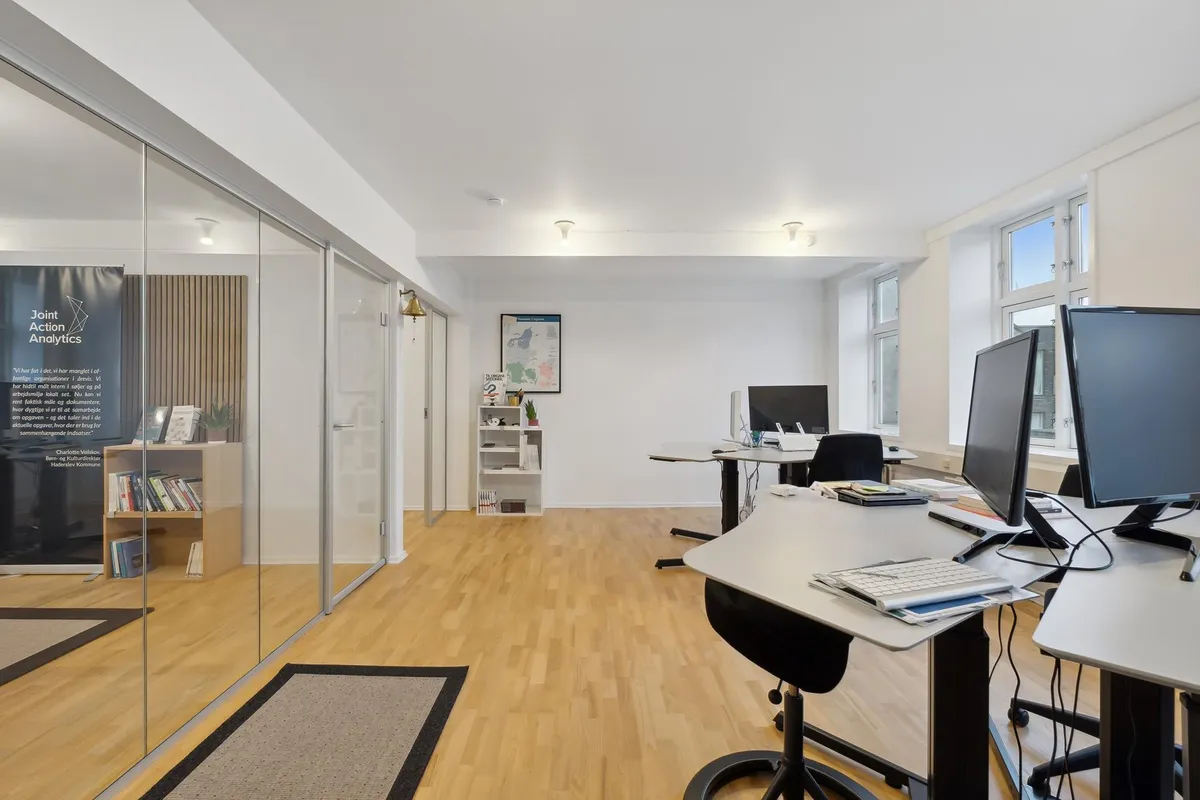 En moderne kontor med højt loft, to skriveborde, to ståborde, lyse gulve og hvide vægge. Der er en åben dør og vinduer, hvor