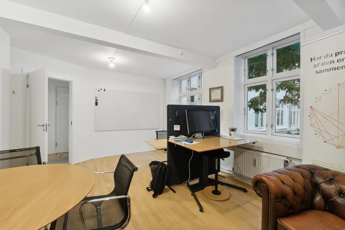 Kontor med lyse gulve, moderne møbler, to skriveborde, tre stolpe, højtalere, computere, lysegardiner og hængevifte