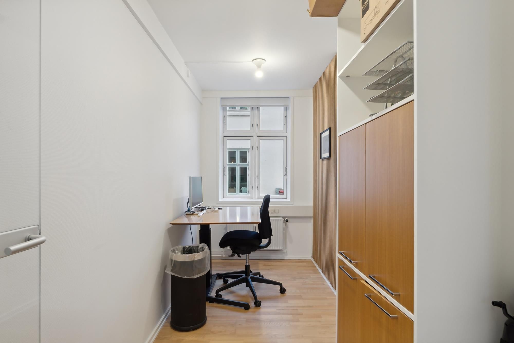 En lille kontorplads med skrivebord, stol, computer og skraldspand. En hvid væg med åbent vindue og hylde med tomme hylder.