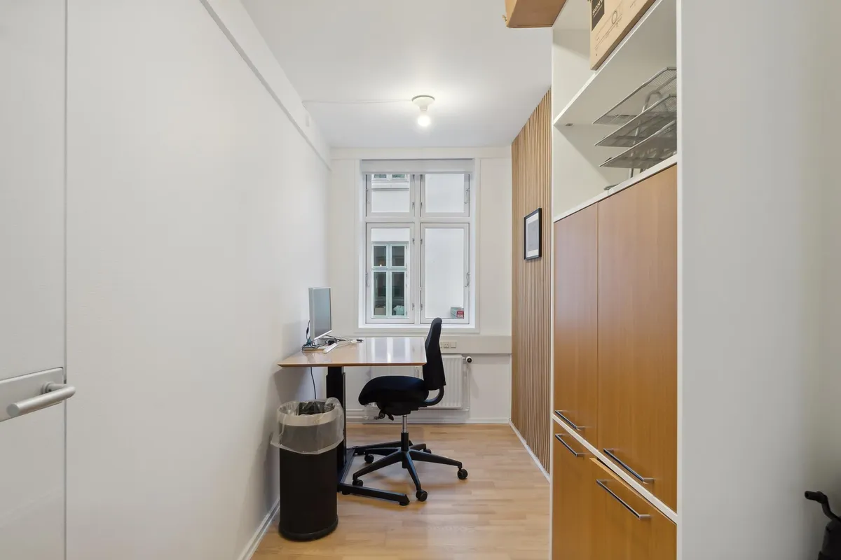 En lille kontorplads med skrivebord, stol, computer og skraldspand. En hvid væg med åbent vindue og hylde med tomme hylder.