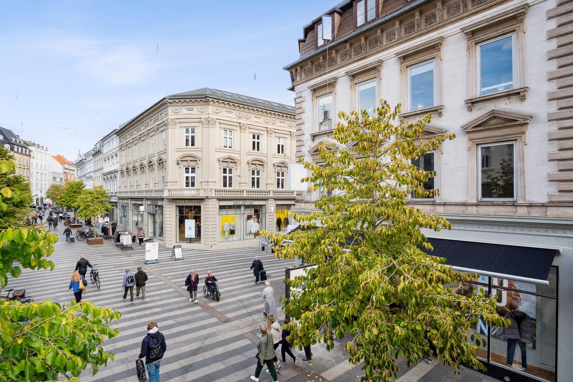 Mennesker går rundt og søger forretninger i en gågade med mange forretninger, en gårdhave og en stor træ i midten