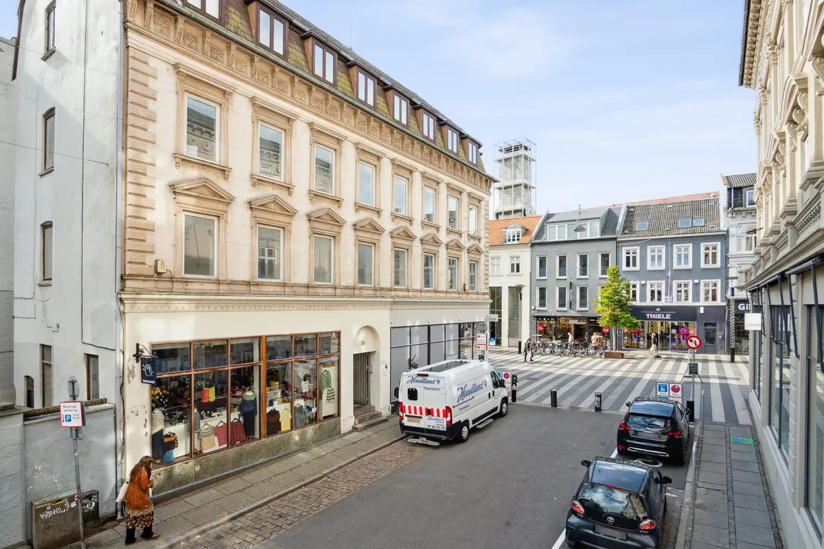 Huse med forretninger og biler parkeret på en gade med en kvinde, der går og kigger i butikken