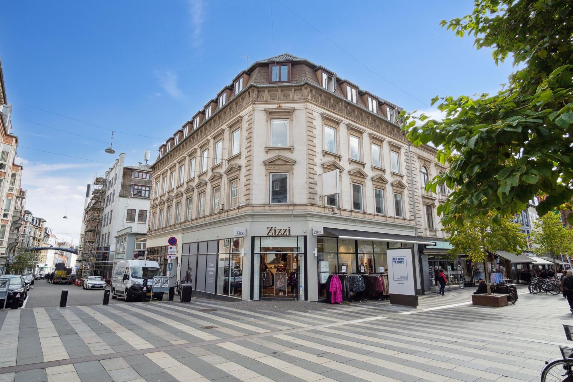 Et moderne tværgående hus med butikker og parkeringspladser. En hvid lastbil står parkeret på en zebraoverskæring. Mennesker