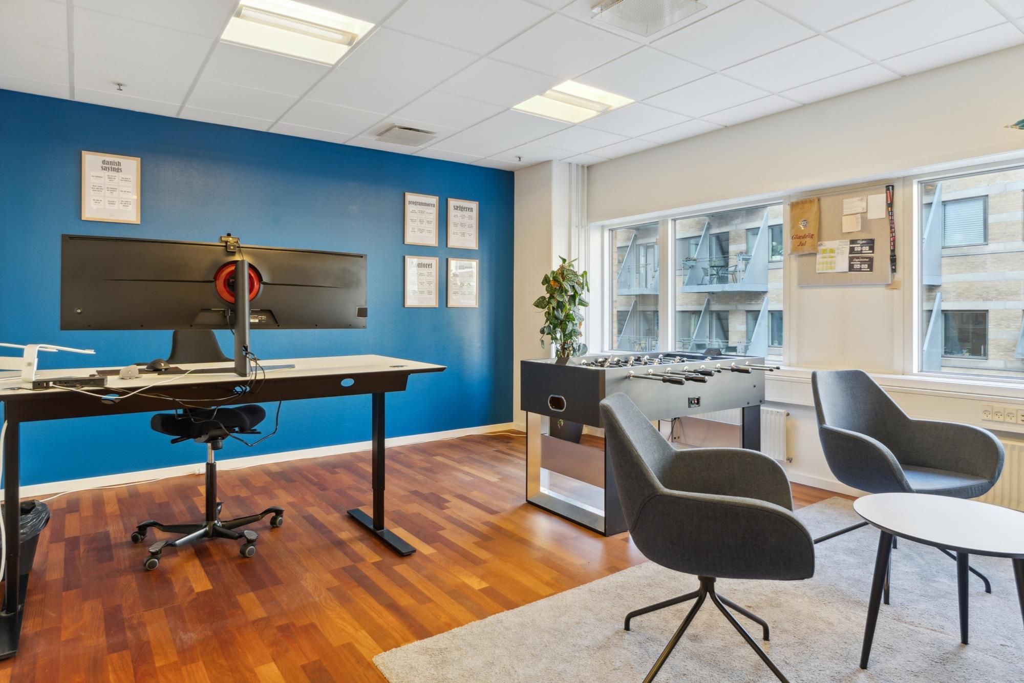 Lyst og åbent kontor med en stor skærm, skrivebord, stol og fodboldbord samt plante og møbler i en moderne indretning