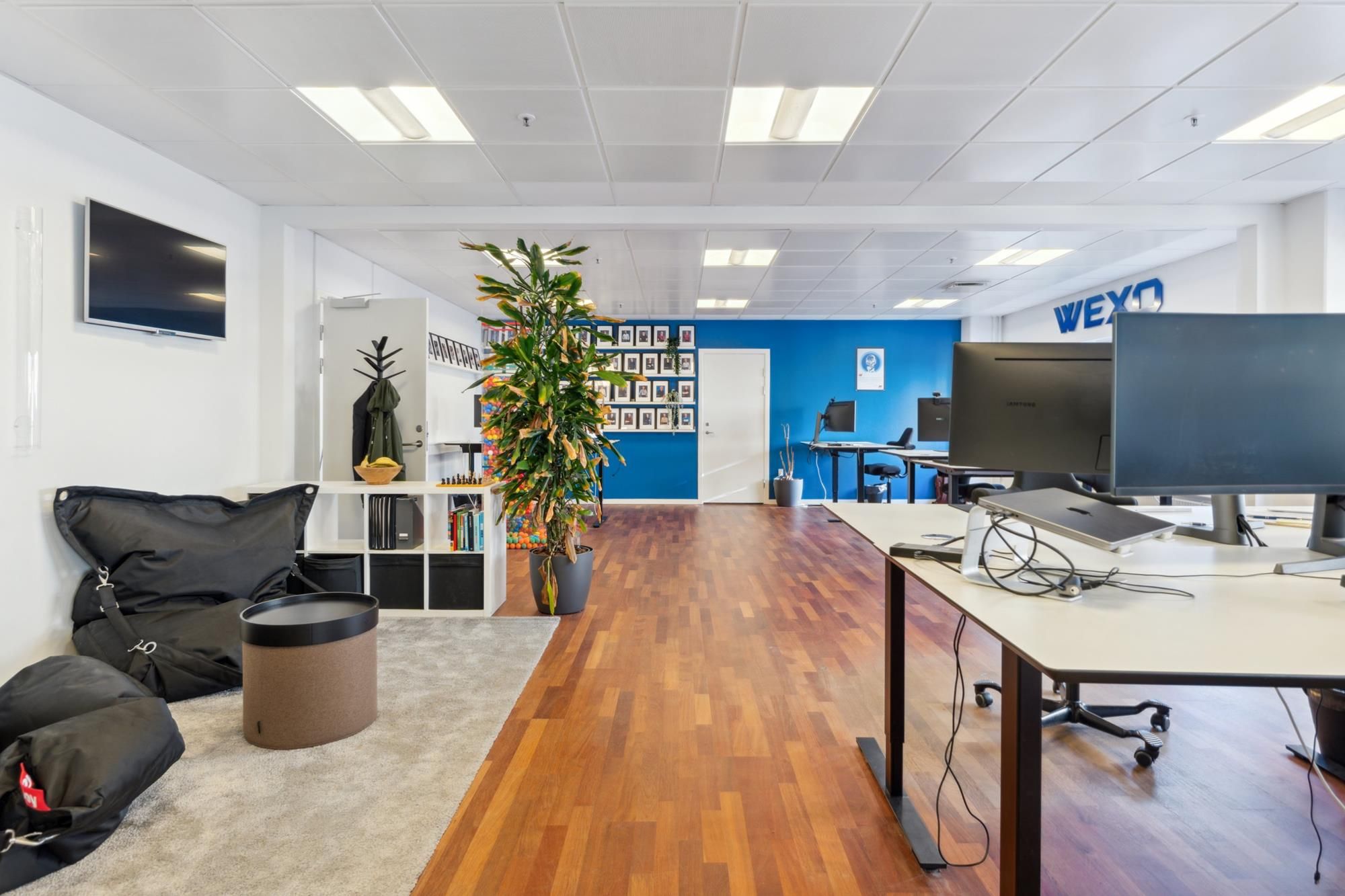 Lyst kontor med højt til loftet, tv og ståborde, lysegule gulvfliser, loftsbelægning, plante og rekreative sofaer i et hjørne