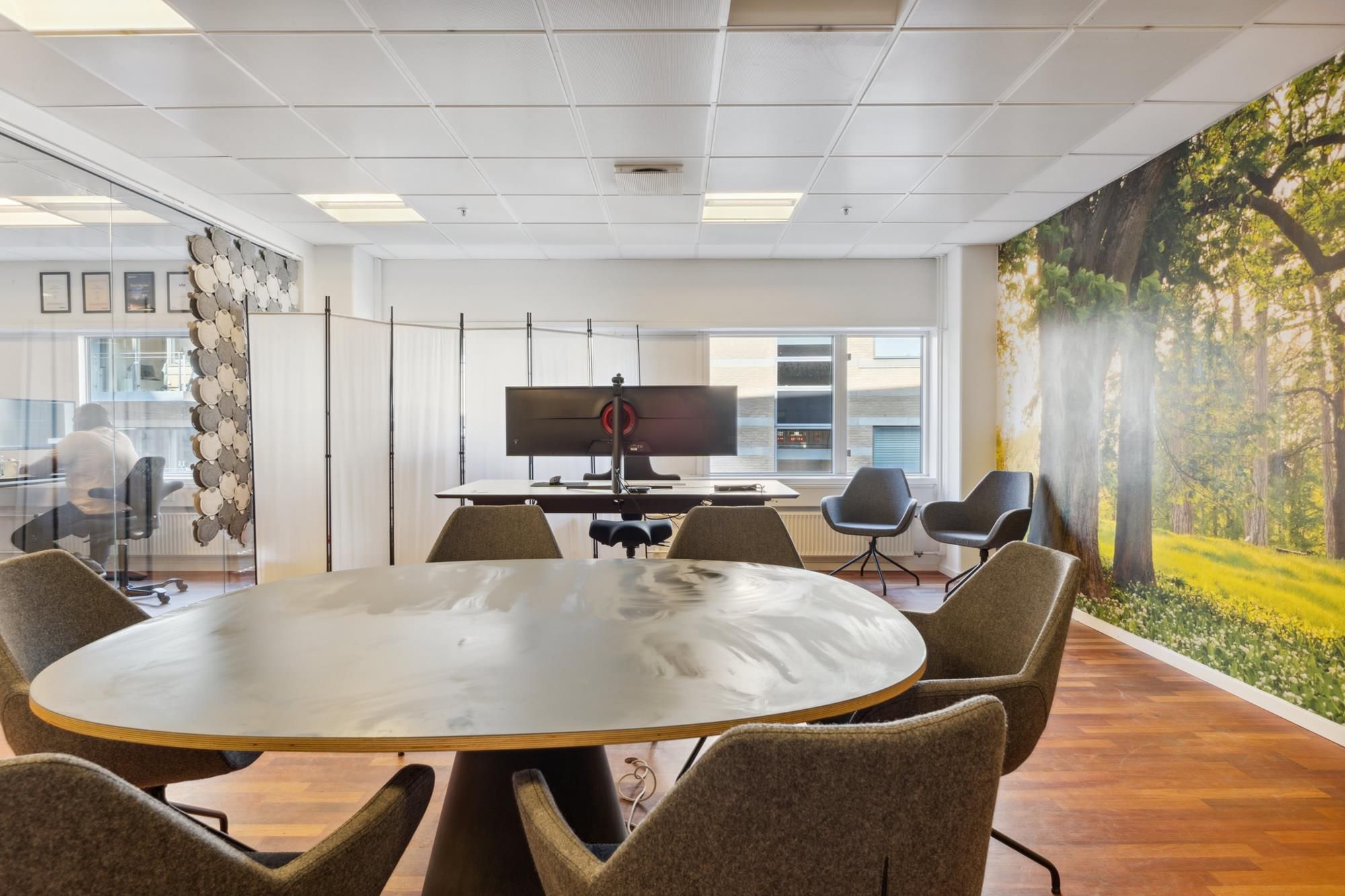 Kontor med moderne møbler, højt loft og kunstværk på væggen med skovbillede og lyse gulve