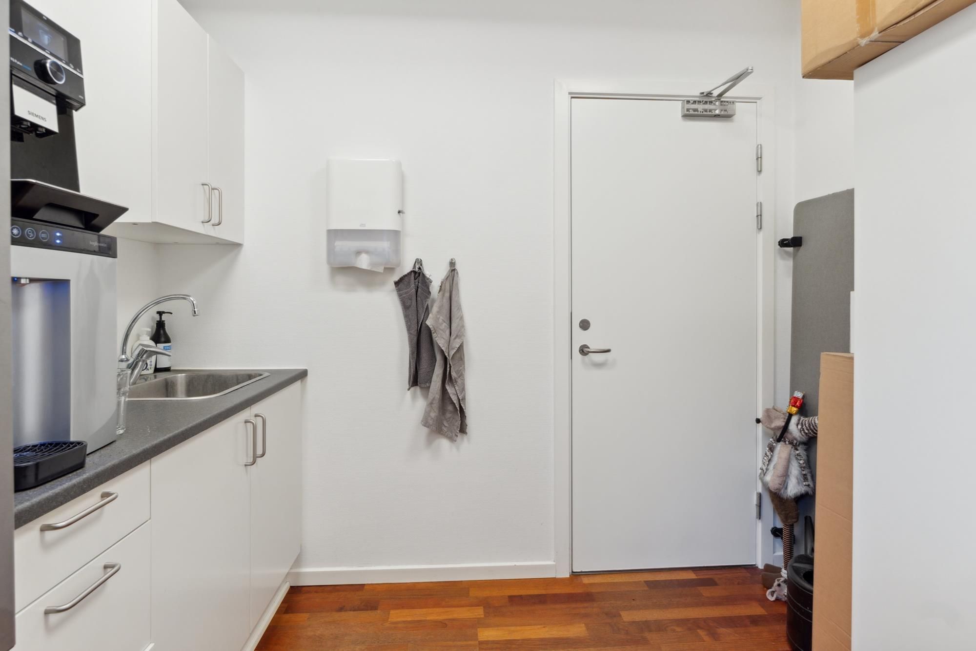 Et lille kontor med en kaffemaskine, en håndvask og to hængede tøjstykker på væggen med en åben dør til siden af lokalet