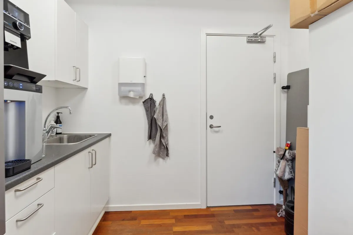 Et lille kontor med en kaffemaskine, en håndvask og to hængede tøjstykker på væggen med en åben dør til siden af lokalet