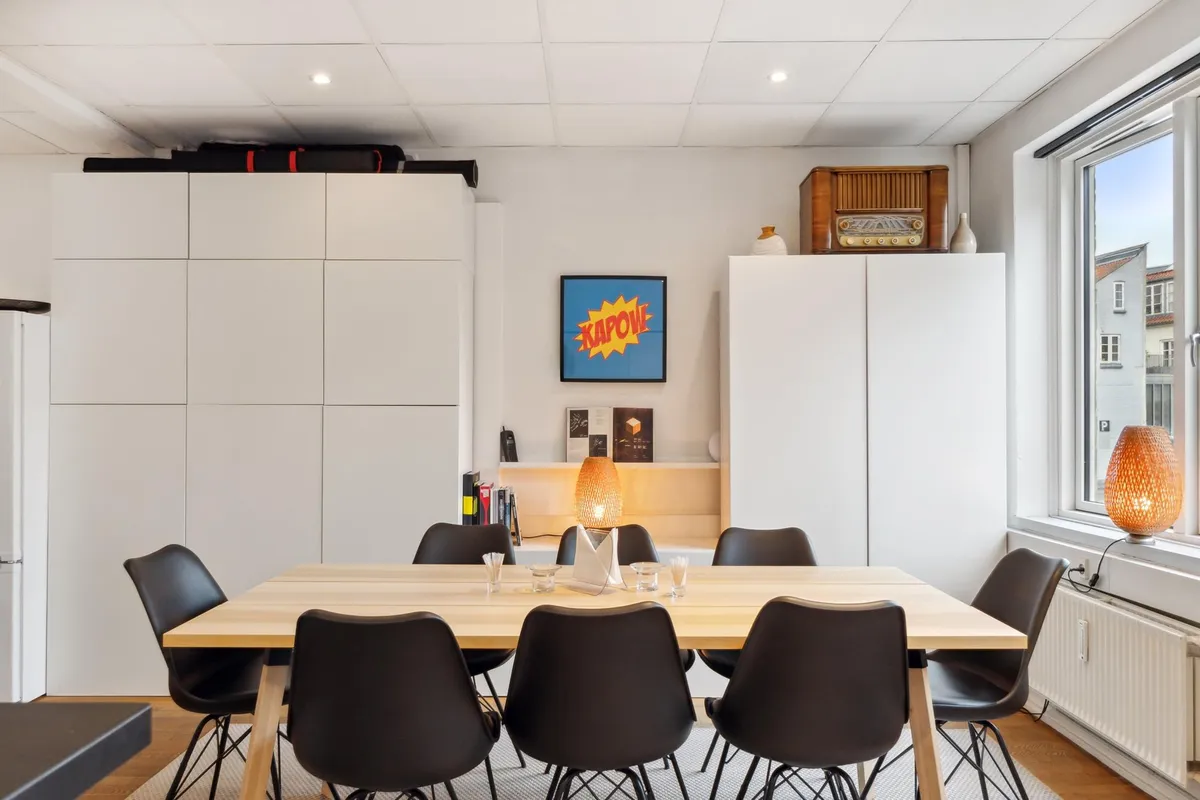 Lyst og moderne køkken med højloft og lyse lamper samt bord og stole samt flotte møbler og loftsrum med kommoder og vindue